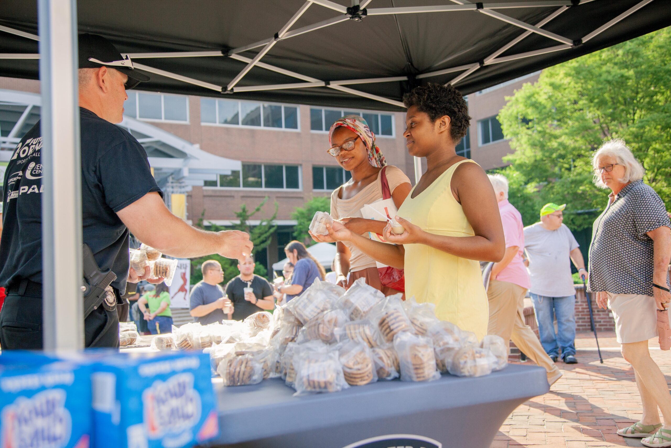 Two women interact with a vendor at a food stand, selecting cookies packaged in clear bags, while a crowd enjoys the event in the background. The scene captures a vibrant community gathering with a focus on food and social interaction.