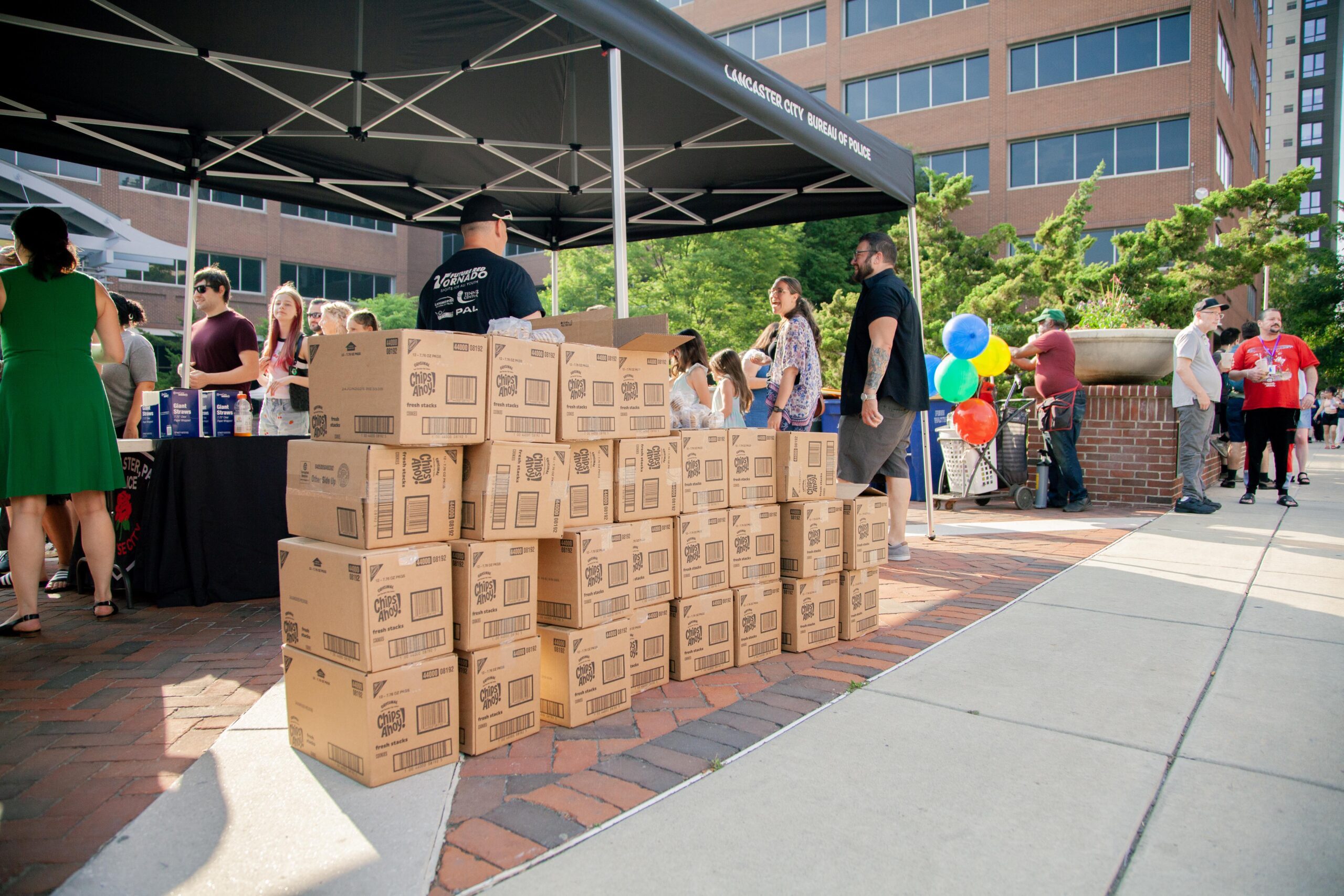 Community event featuring a crowd gathered around a tent with the Lancaster City Bureau of Police, showcasing stacked boxes of snacks and drinks, as attendees interact and enjoy the festivities.