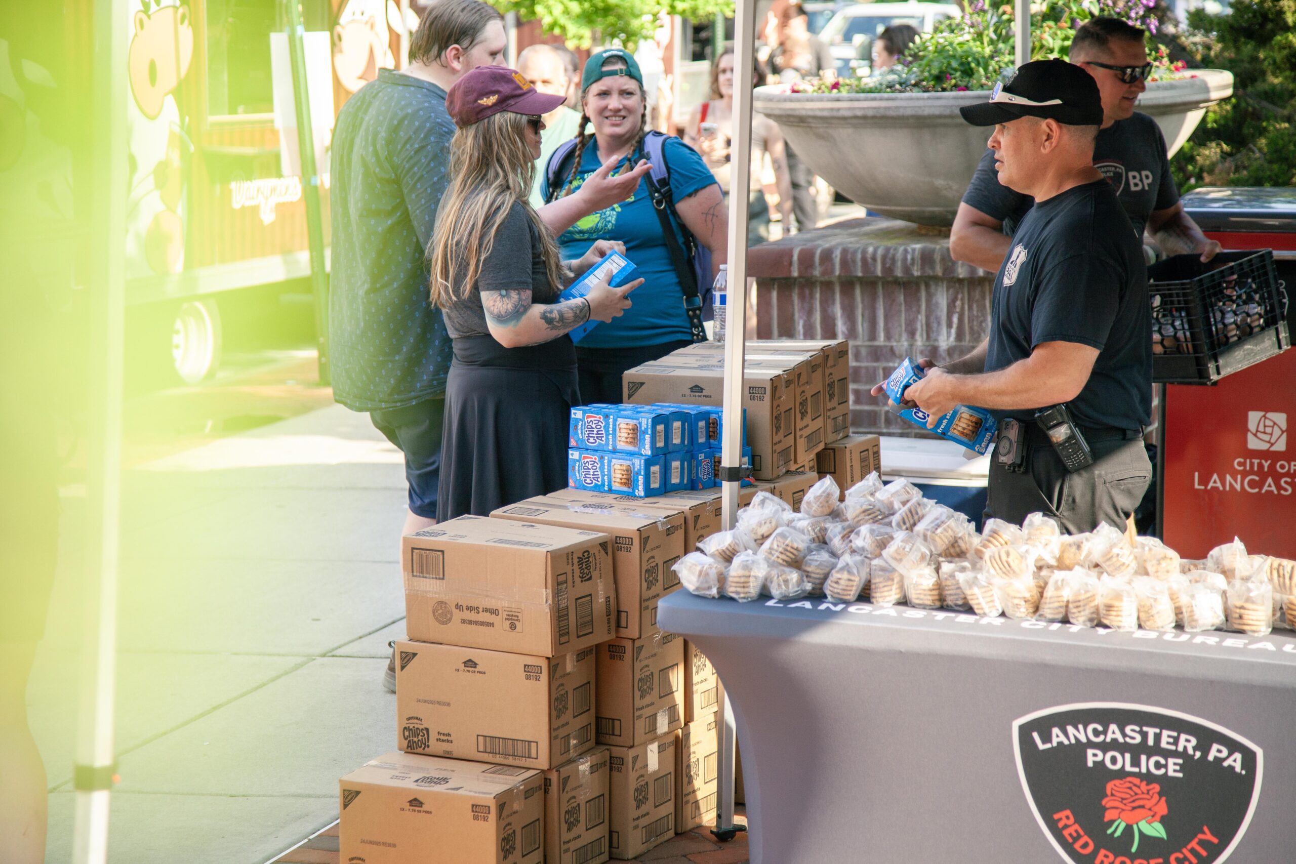 A community event in Lancaster, PA, featuring local vendors and police officers engaging with attendees. Boxes of snacks and baked goods are displayed on tables, while people converse and enjoy the atmosphere.