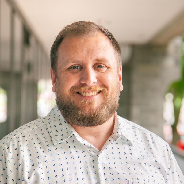 Smiling man with a beard wearing a white shirt with blue patterns, standing in a modern outdoor corridor with greenery in the background.