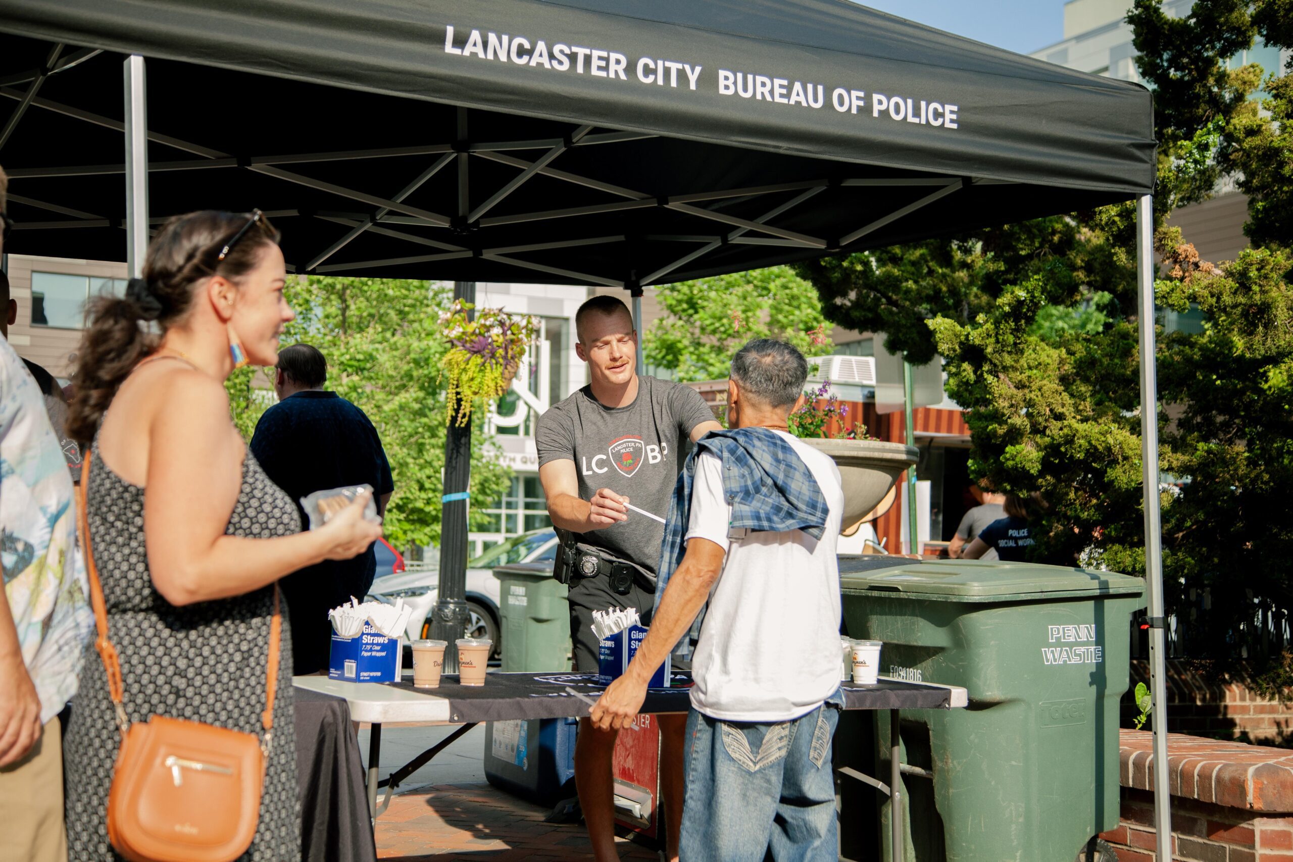 Lancaster City Bureau of Police community outreach event, featuring a police officer engaging with local residents at a table with informational materials and coffee cups, set against a backdrop of greenery and urban architecture.