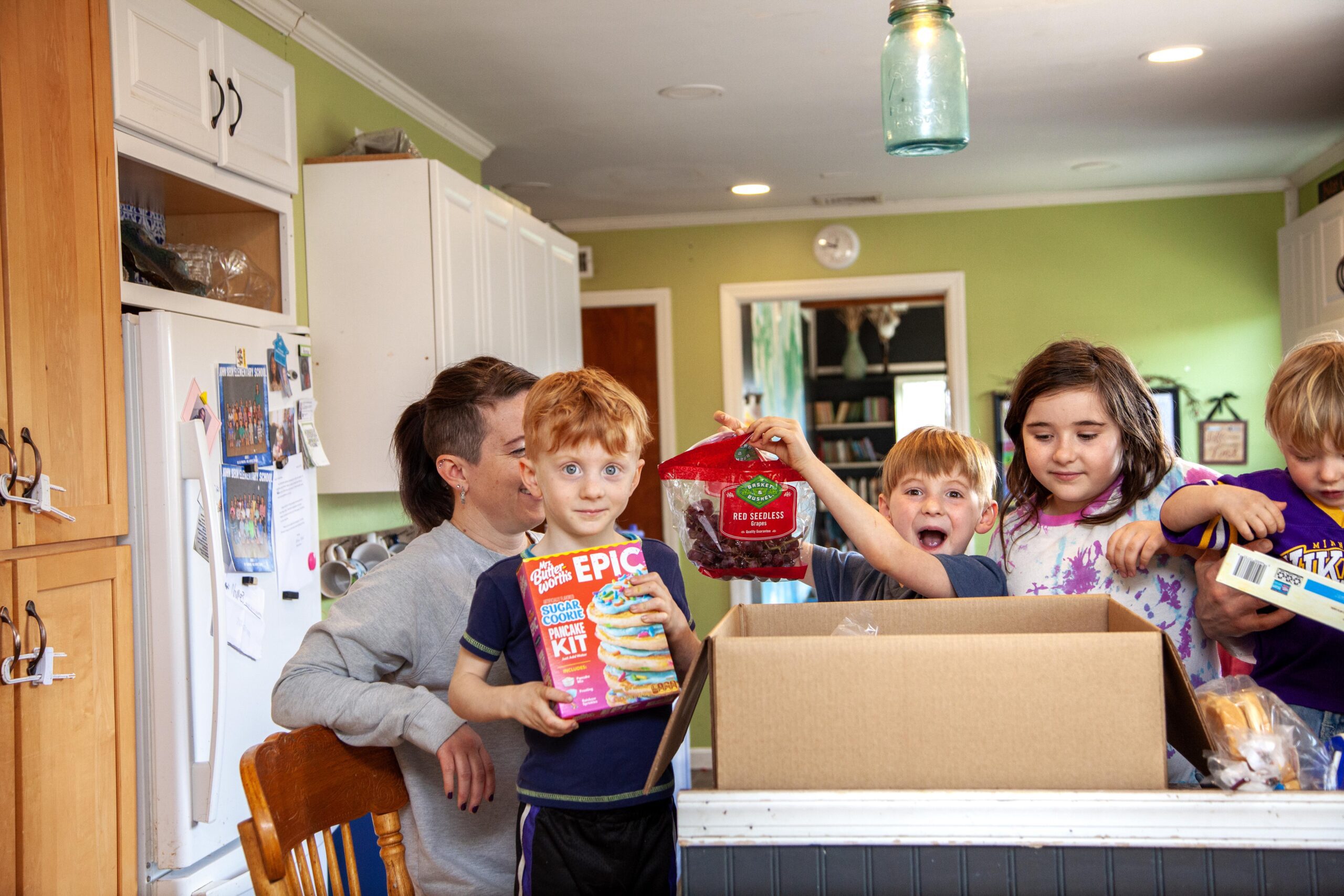 Children excitedly unpack a box in a bright kitchen, holding a pancake kit and a bag of red seedless grapes, while an adult smiles in the background.