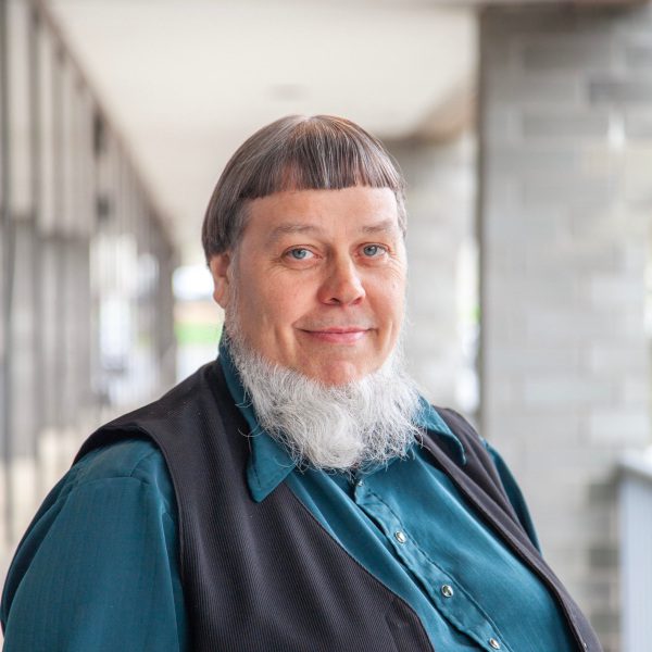 Portrait of a person with a unique hairstyle and a full beard, wearing a blue shirt and black vest, standing in a modern architectural setting with blurred background elements.