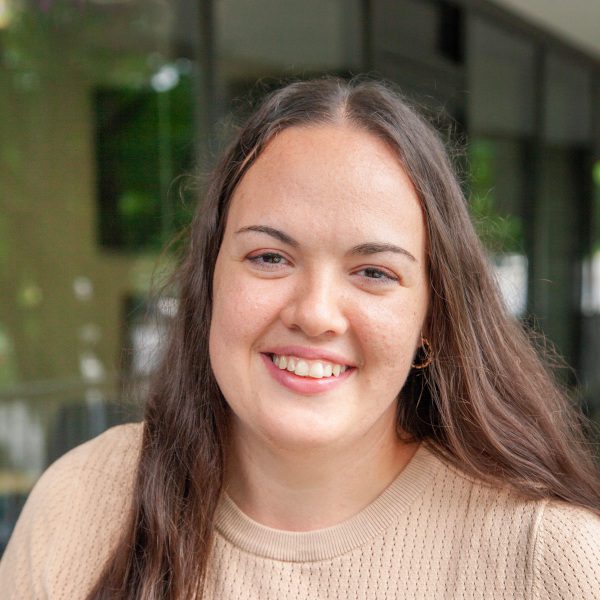 Smiling woman with long brown hair wearing a beige sweater, standing outdoors in front of a glass building, representing a friendly and approachable demeanor.