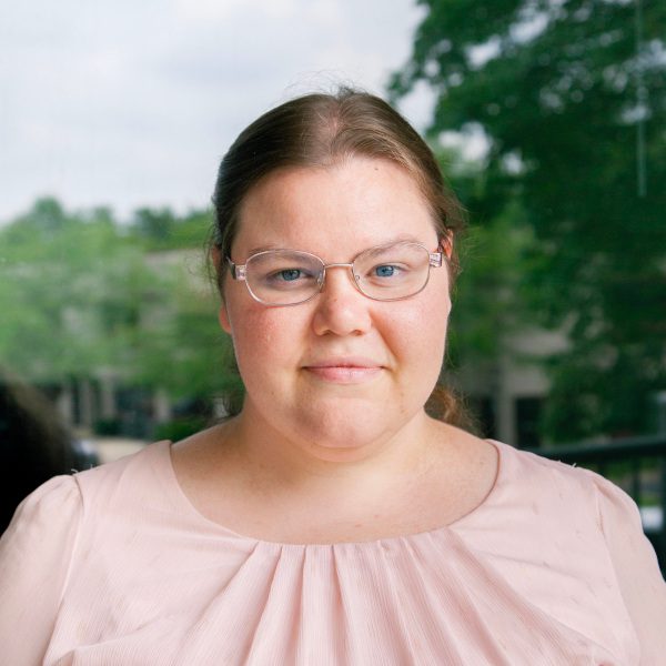 Portrait of a smiling woman with glasses, wearing a light pink blouse, standing in front of a window with greenery in the background. The image highlights a professional atmosphere.