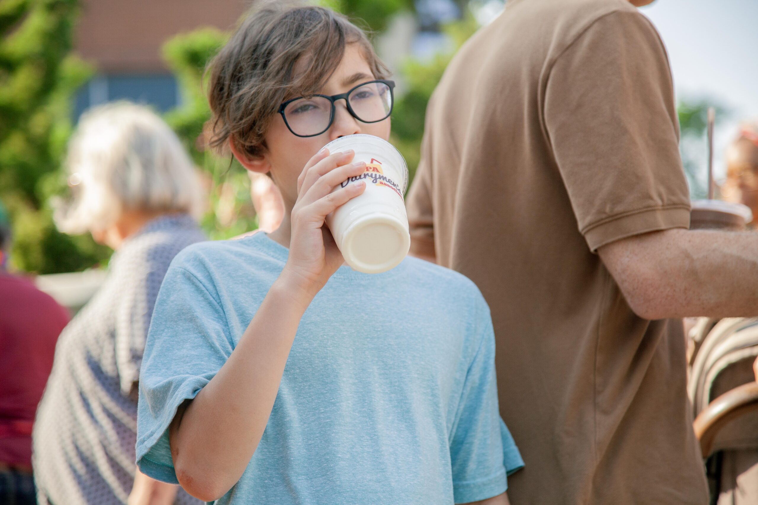 Child enjoying a beverage in a busy outdoor setting, wearing a light blue shirt and glasses, with people mingling in the background.