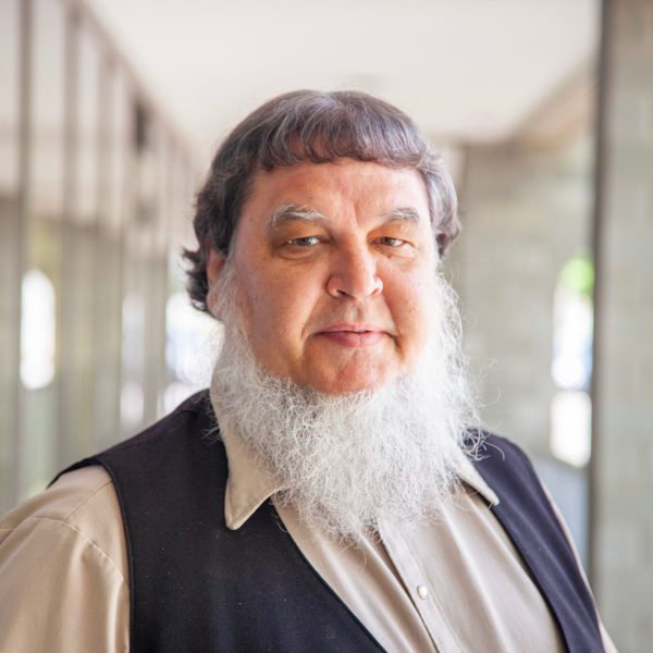 Portrait of a middle-aged man with a long beard and short, wavy hair, wearing a dark vest over a light shirt, standing in an indoor corridor with natural light.