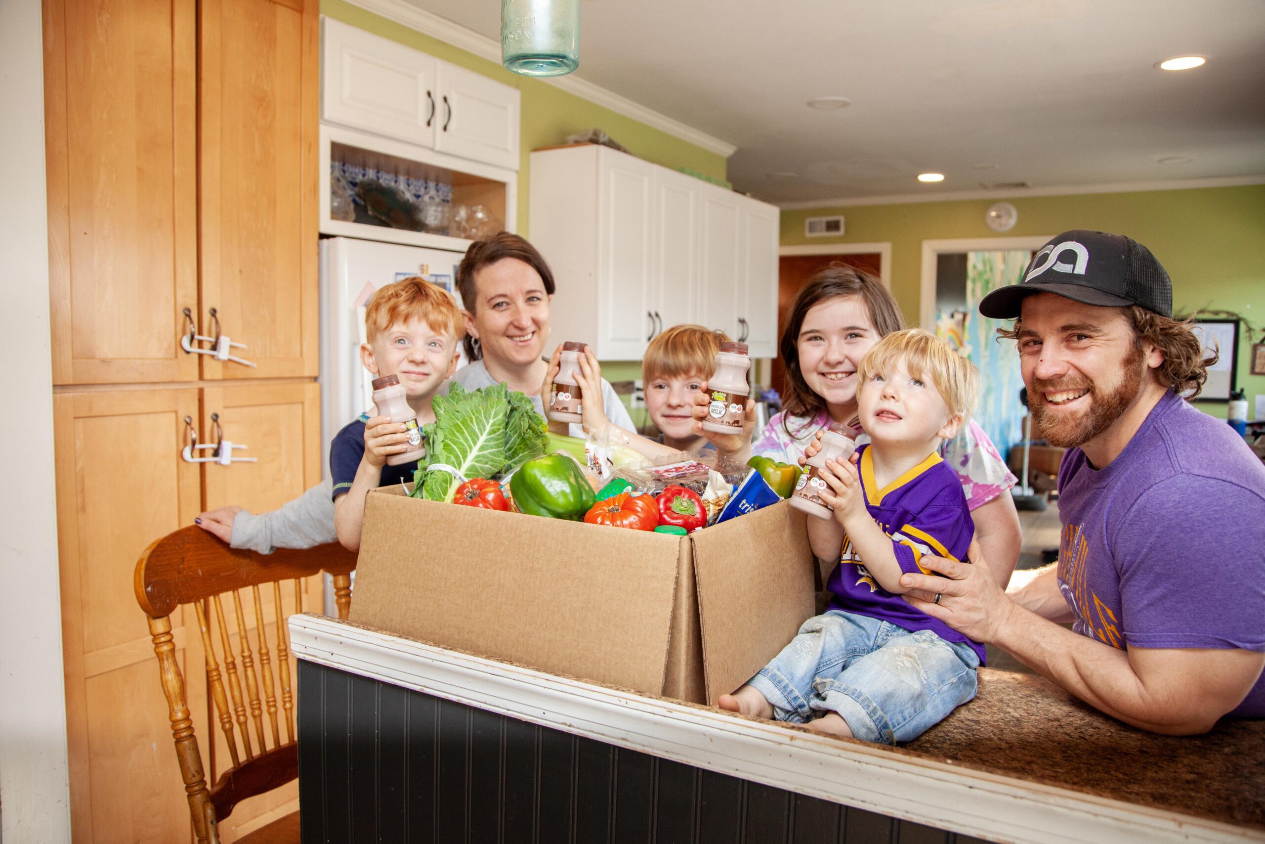 Family enjoying healthy groceries in a kitchen, featuring children holding drinks and a box filled with fresh vegetables like lettuce, bell peppers, and tomatoes.