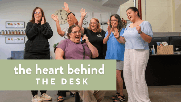Group of smiling women celebrating together in an office setting, with one woman on the phone. The background features a clock and shelves with jars, emphasizing a lively and supportive work environment.