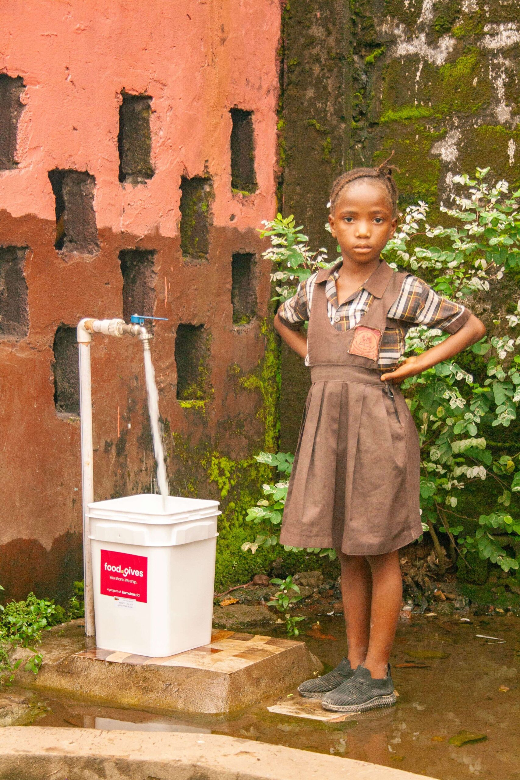 Young girl in a brown dress stands beside a water tap, with a white bucket labeled "food.gives" collecting flowing water. The background features a textured pink wall and greenery, highlighting the importance of clean water access in the community.