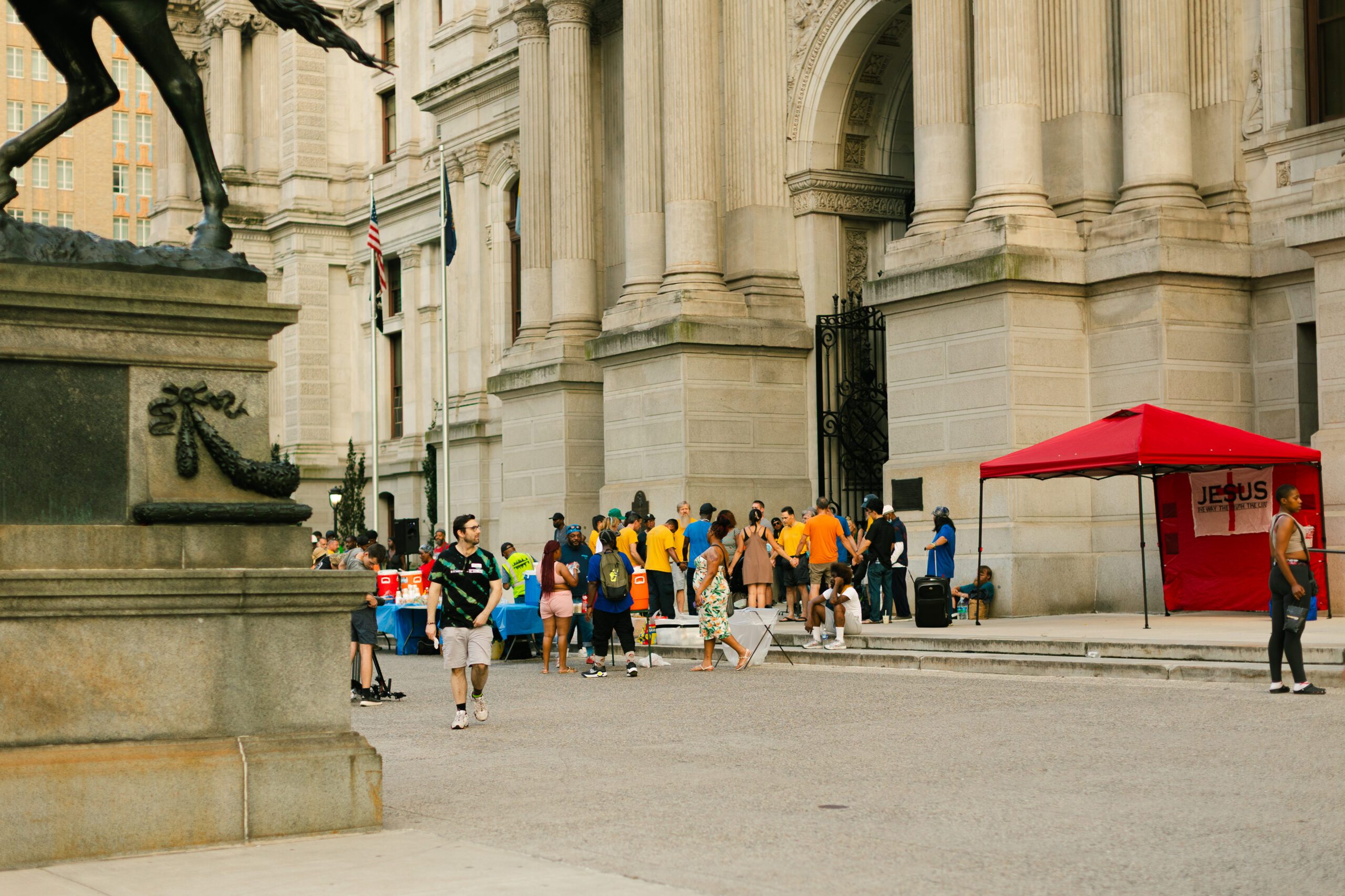 Crowd gathered outside a historic building, featuring a red tent with "JESUS" signage, as people in colorful shirts engage in activities. A statue of a horse is visible in the foreground, adding to the urban atmosphere.