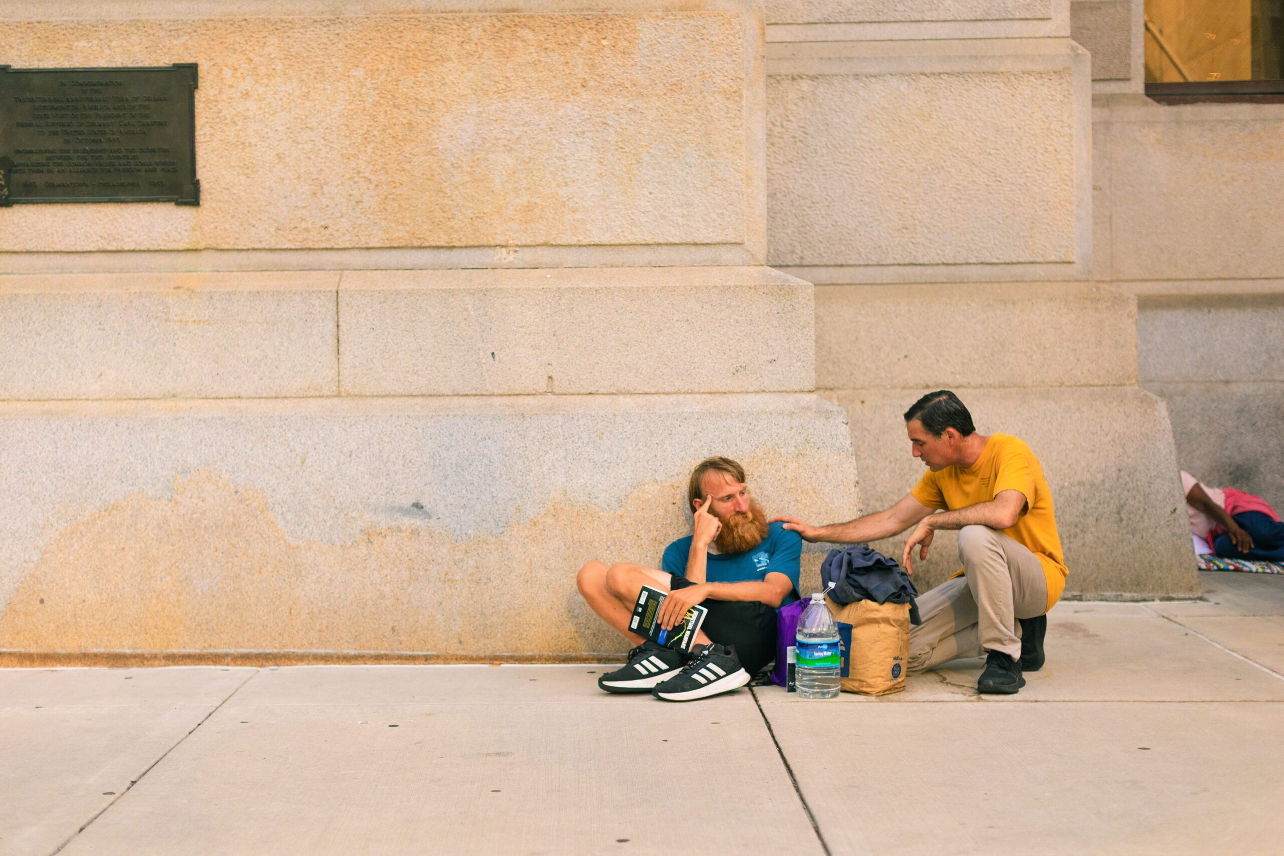 A man with a beard sits on the sidewalk, looking contemplative, while another man in a yellow shirt offers support. They are near a stone wall, surrounded by personal belongings and water bottles, illustrating a moment of connection in an urban setting.