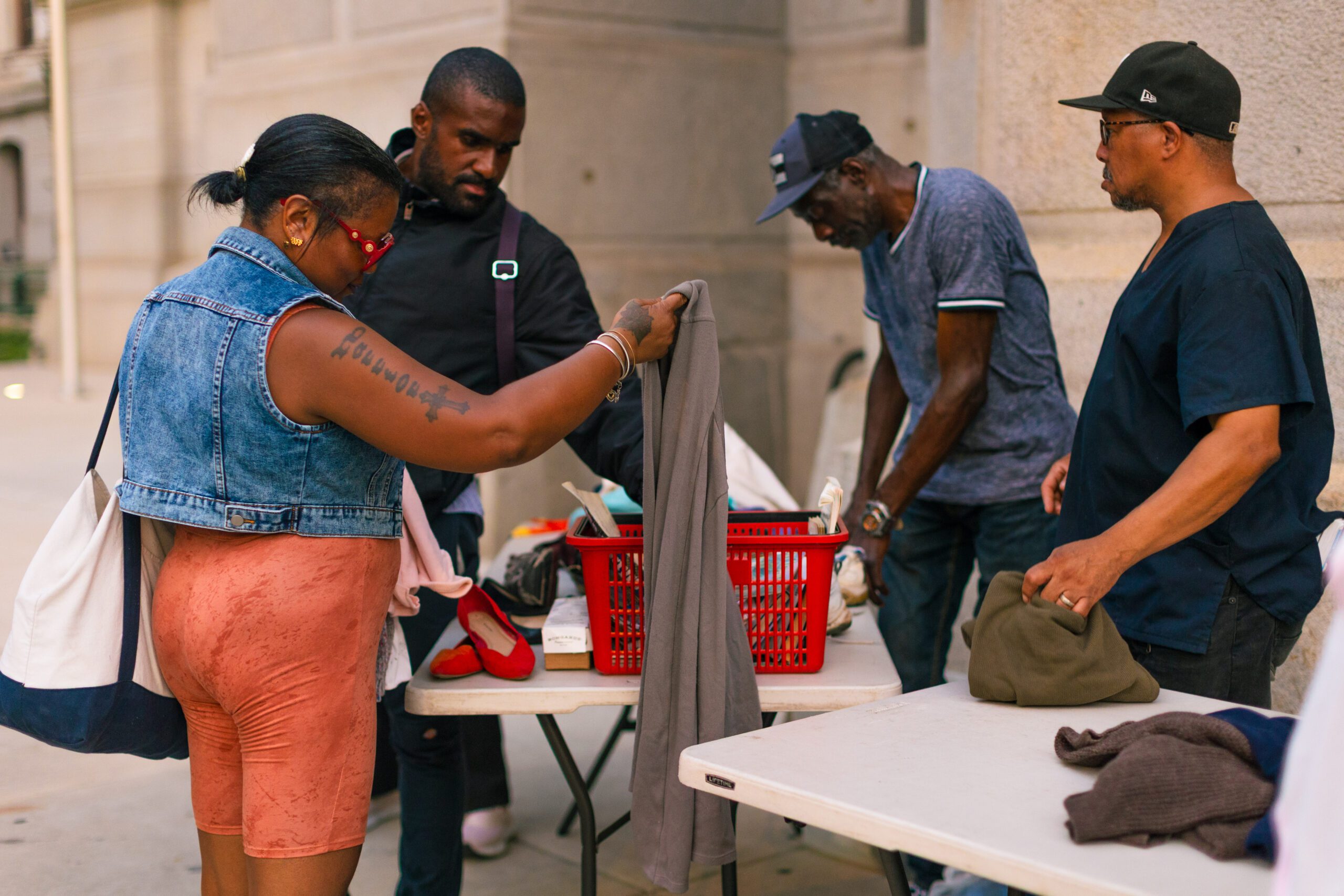 A group of four individuals engaging in a community clothing exchange, with one woman examining a piece of clothing while others sort through items on a table, showcasing a vibrant atmosphere of sharing and support in an urban setting.