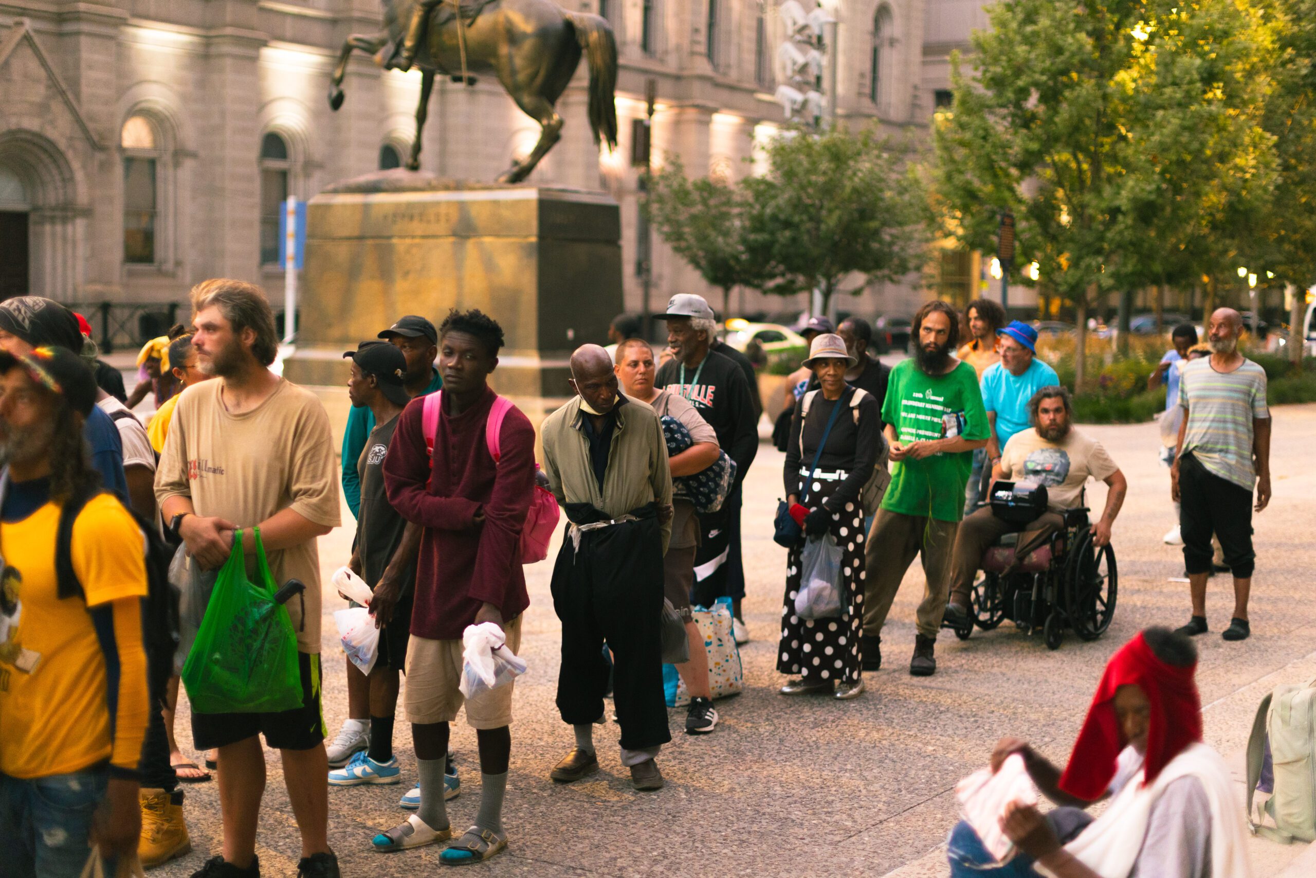 A diverse group of individuals, including men and women of various ages, standing in line outside a historic building, with a statue of a horse in the background. Some individuals hold bags, while one person is in a wheelchair, indicating a community gathering or support event.