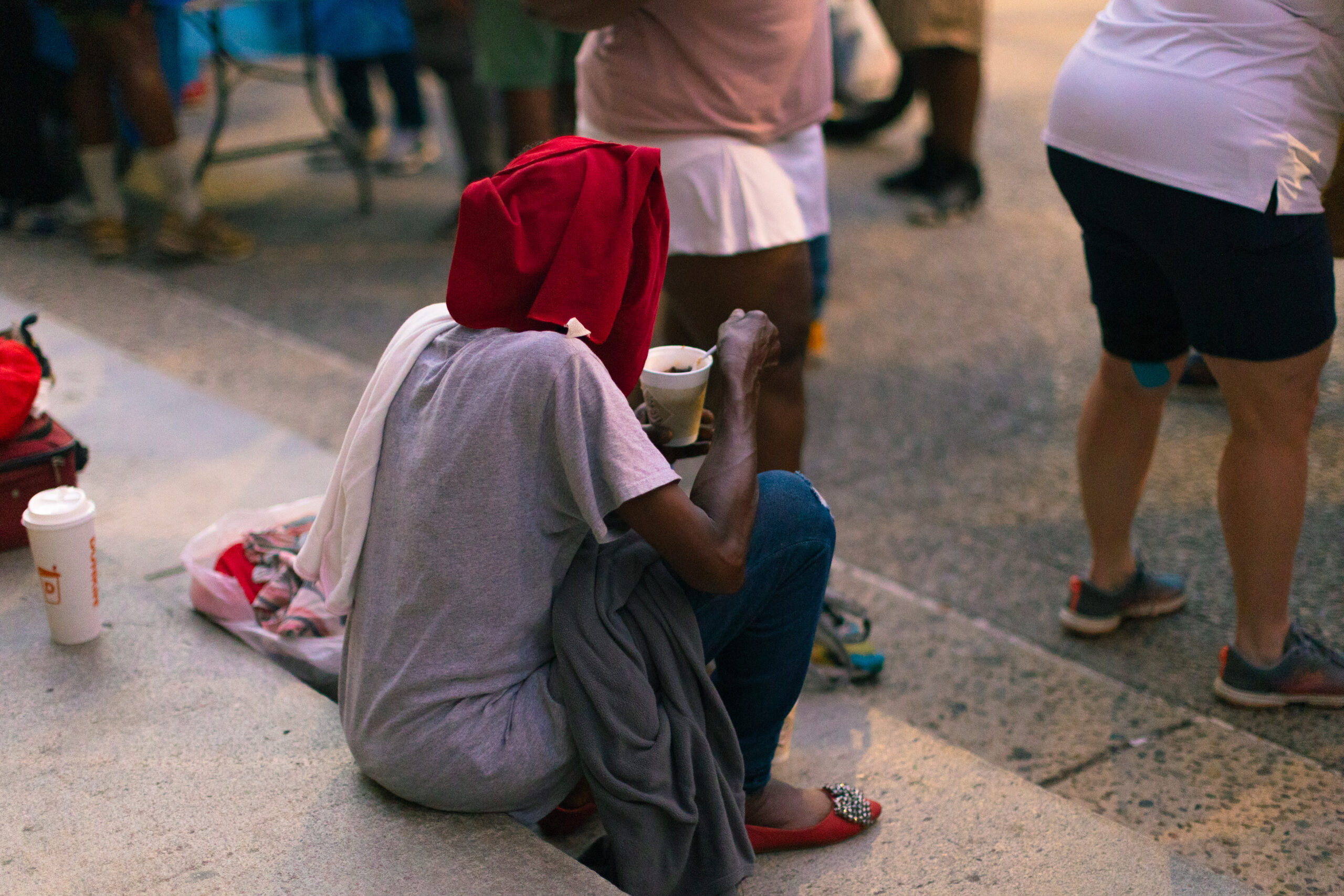 A person wearing a red hood sits on a step, holding a cup and looking down, surrounded by a crowd of people in a public space. The scene captures a moment of solitude amidst the busyness of the urban environment.