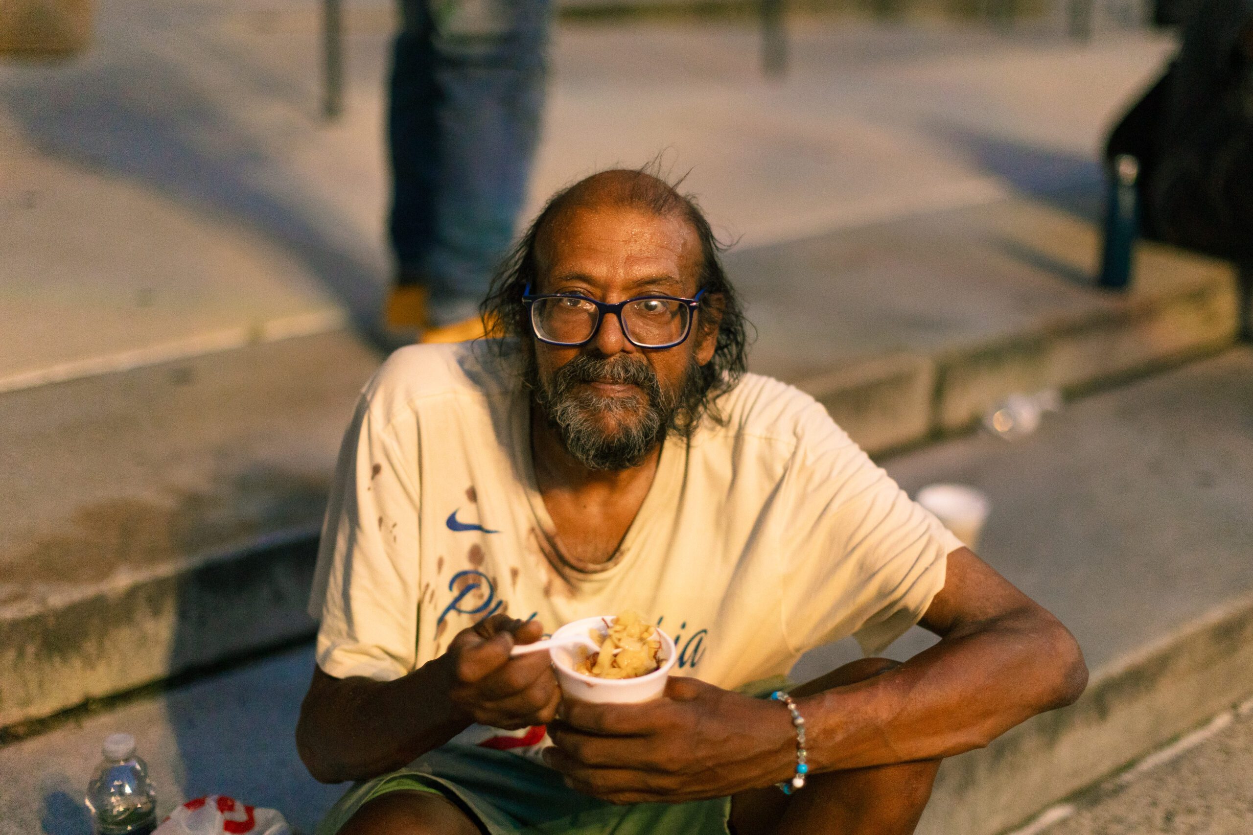 A homeless man sitting on steps, holding a cup of food, with a weary expression. He has long hair, glasses, and is wearing a stained t-shirt. Nearby, there are water bottles and other discarded items, highlighting urban poverty.