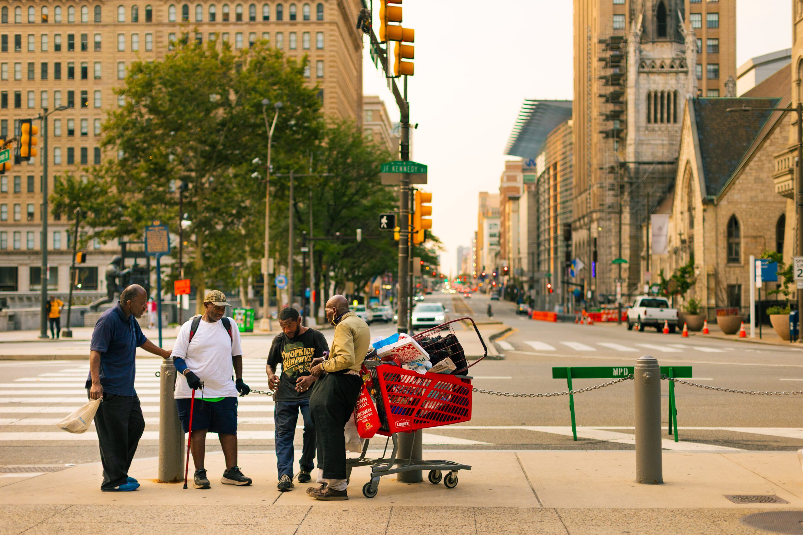 Group of four men gathered on a city sidewalk, discussing and examining items in a shopping cart. Urban setting with traffic lights and buildings in the background, showcasing community interactions and street life.