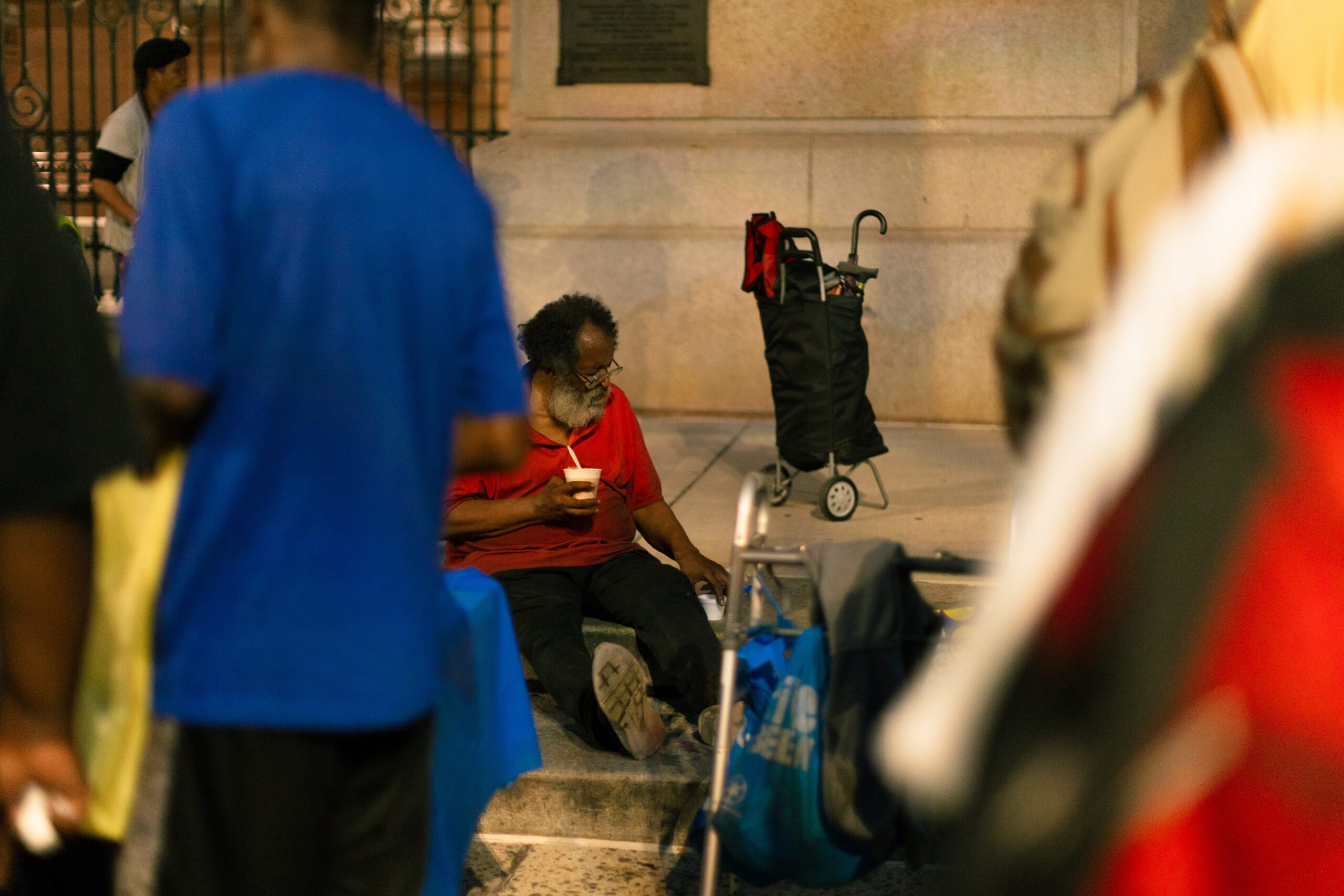 An elderly man with a beard sits on the sidewalk, wearing a red shirt and holding a cup, surrounded by people in a busy urban setting. A shopping cart is nearby, indicating a moment of pause in a communal space.