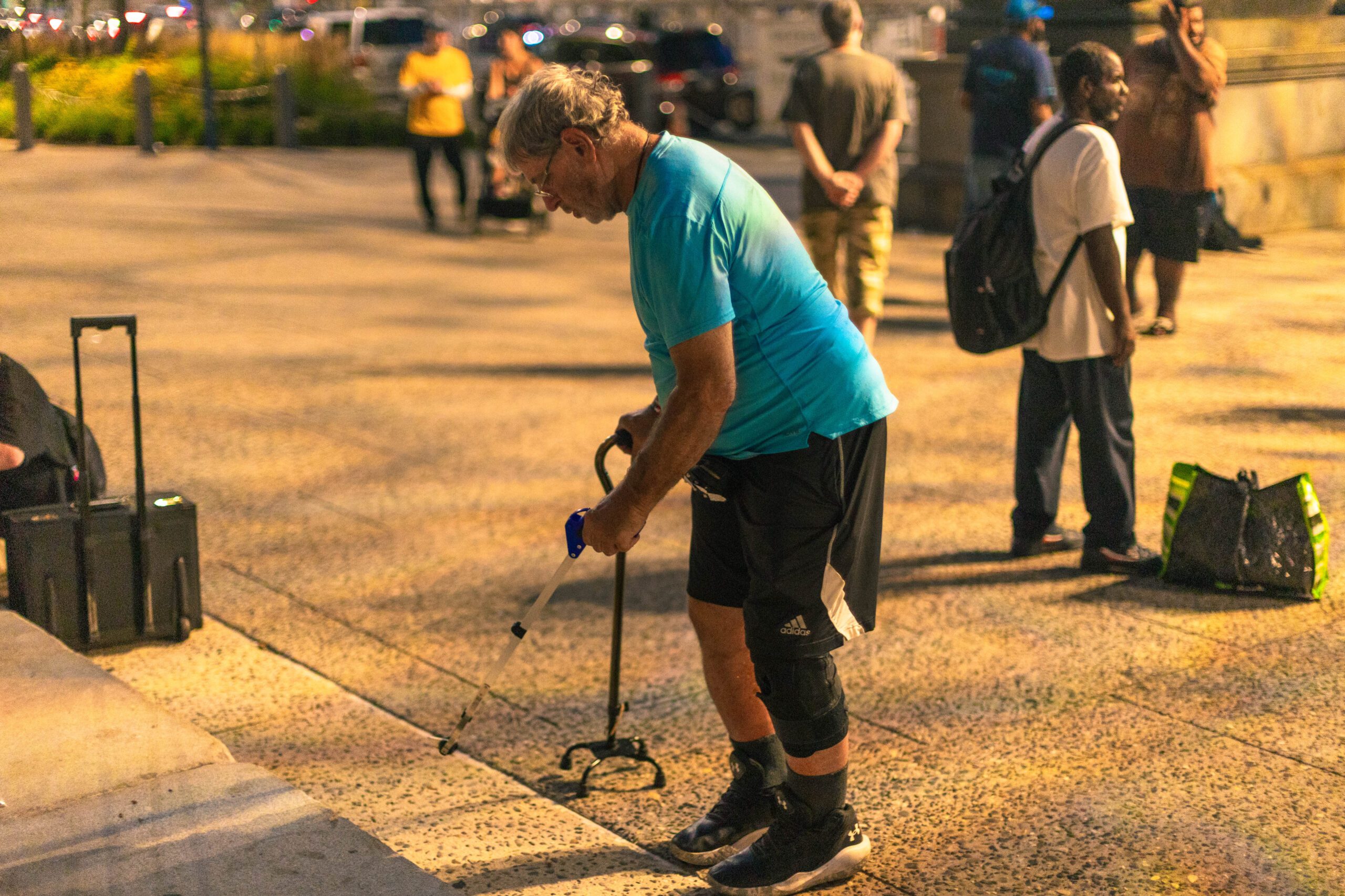 An elderly man in a blue shirt and black shorts uses a grabber tool to pick up items from the ground in a busy urban setting, with other people visible in the background.