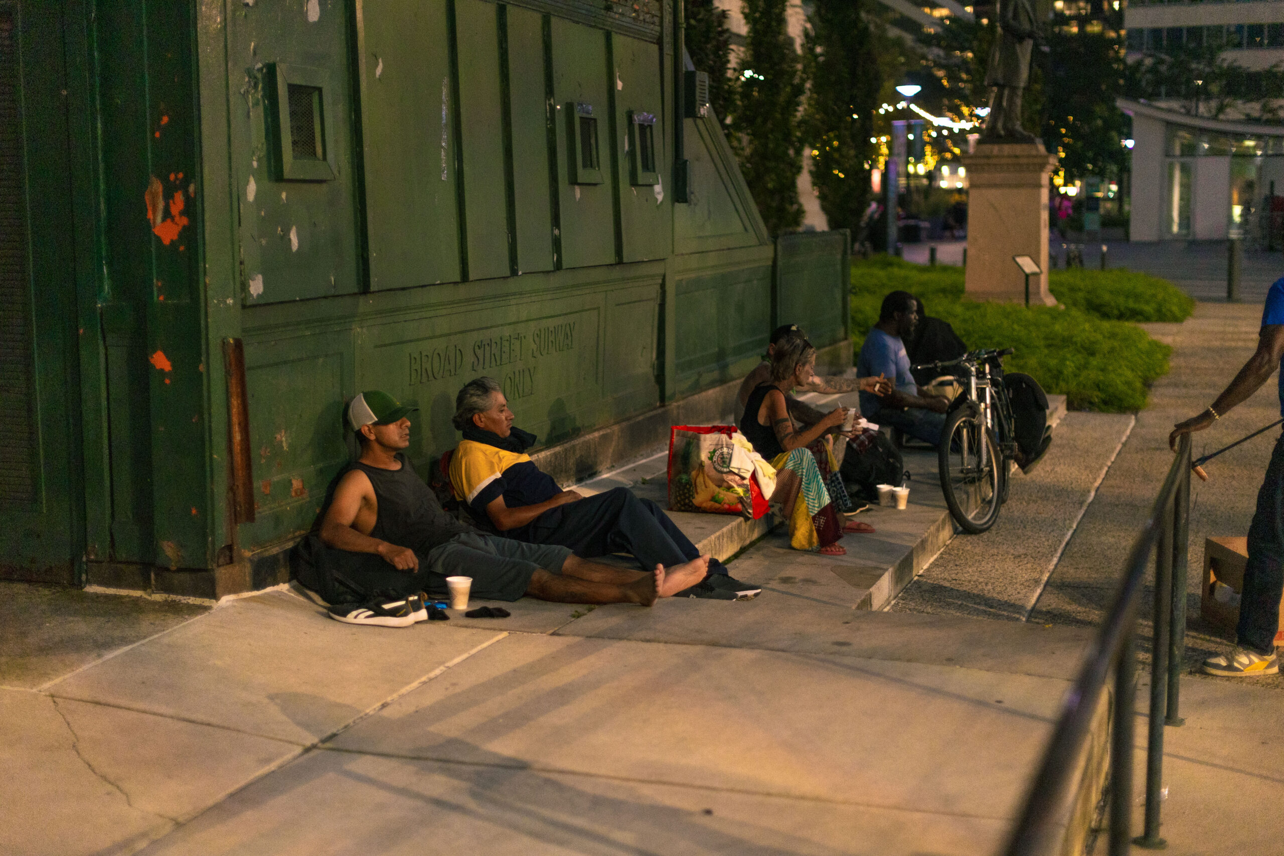 Group of homeless individuals sitting on the steps of a building at night, with some holding cups and bags. The scene captures urban life and the challenges faced by those experiencing homelessness in a city setting.