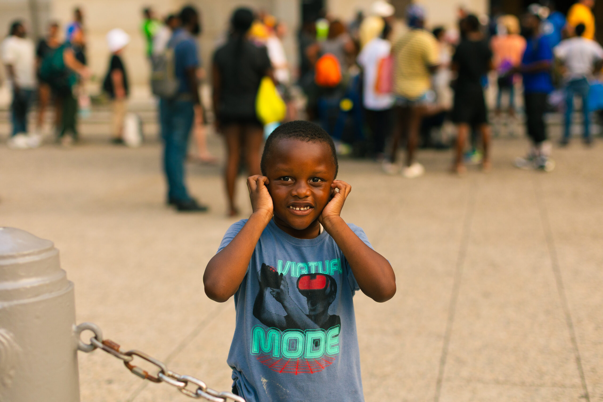 A young boy smiling and covering his ears stands in a busy outdoor setting with a crowd of people in the background, showcasing a lively atmosphere. He wears a blue t-shirt featuring a graphic design, capturing a playful moment amidst the activity.