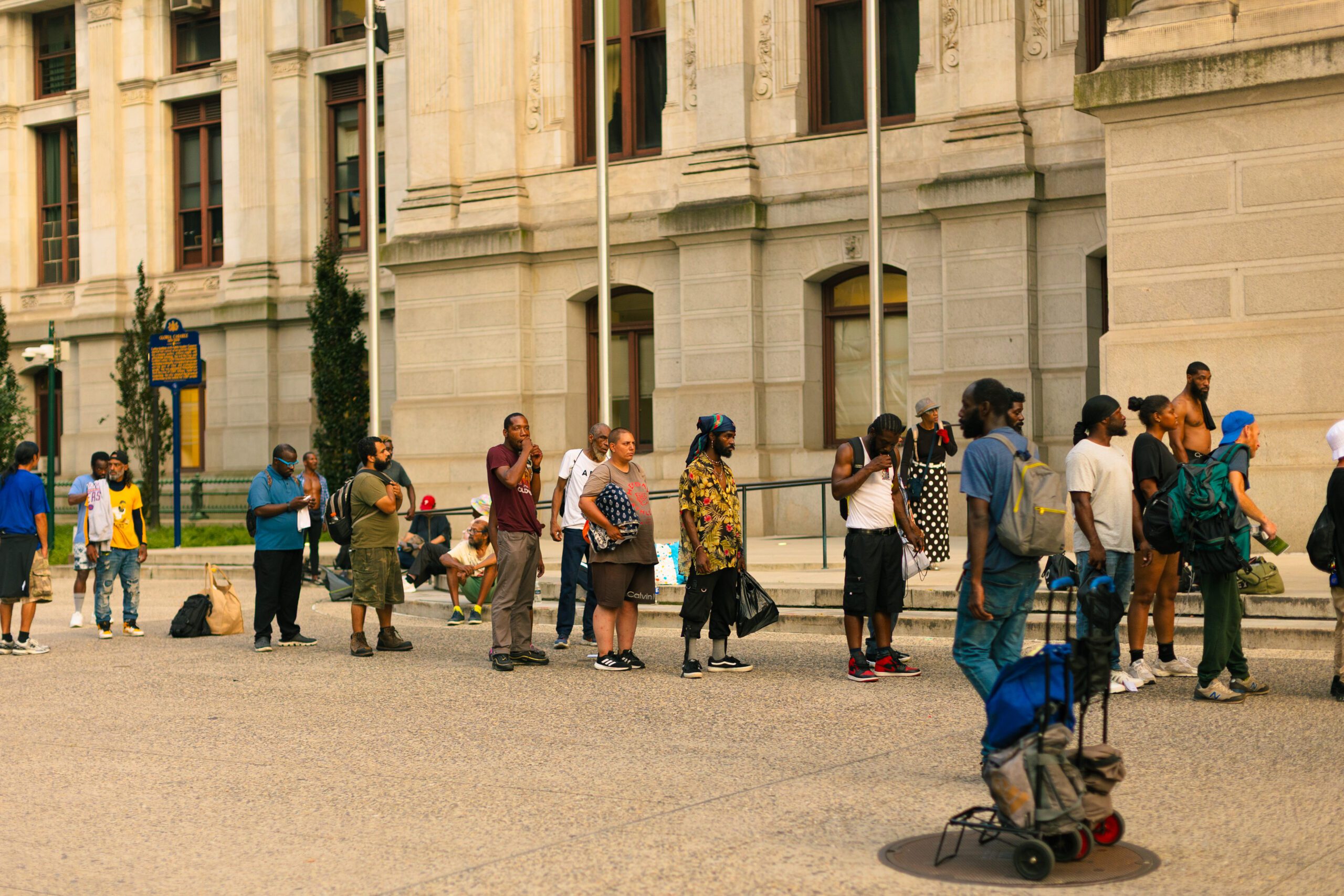 A diverse group of people standing in line outside a historic building, with some individuals engaging in conversation and others using their phones. The scene captures a moment of community interaction in an urban setting, highlighting the social dynamics of public spaces.