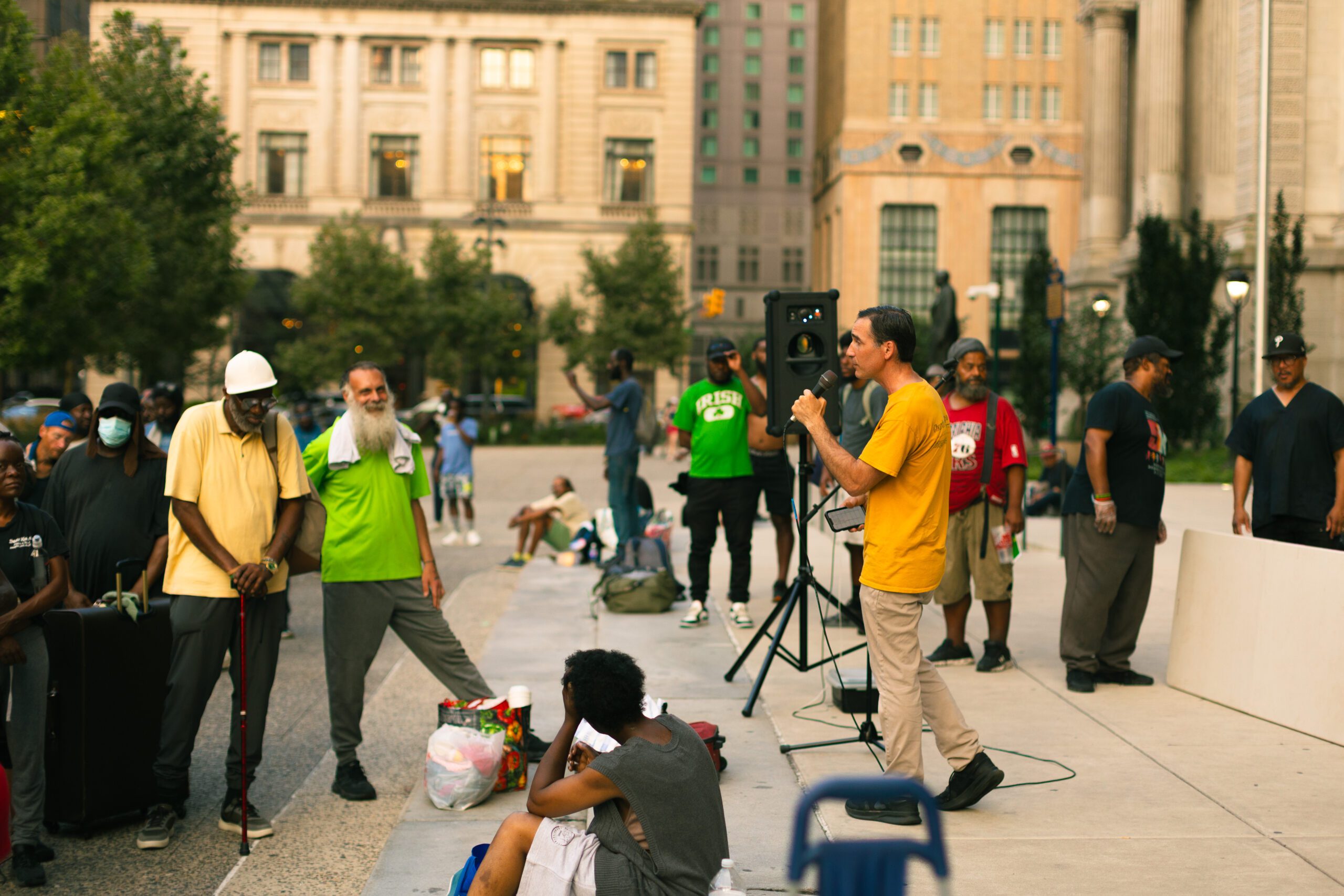 A community gathering in an urban park, with a speaker addressing a diverse audience. Attendees include men and women of various ages, some seated and others standing, listening attentively. The scene captures a moment of engagement and connection, highlighting the importance of community support and activism.