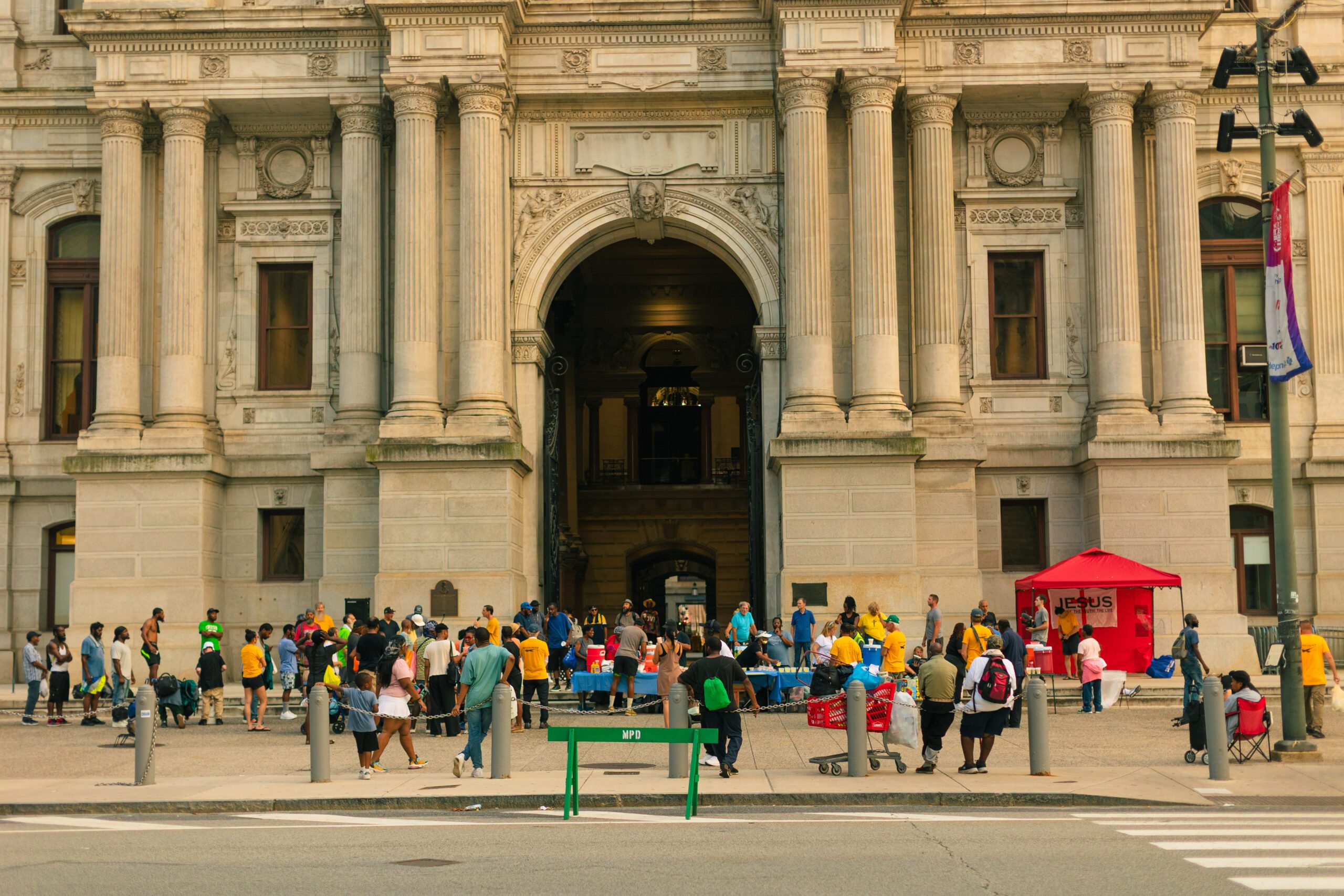 Crowd of people gathered outside a historic building, engaging in community activities and services, with a red tent and tables set up for distribution events.