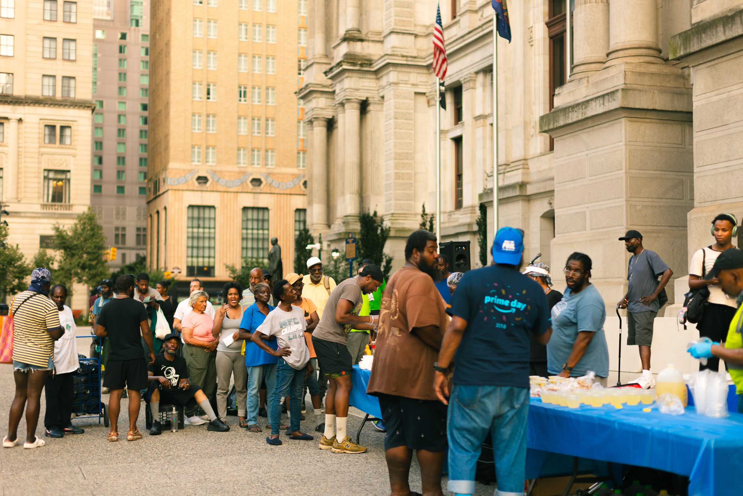 Community members gather outside a historic building for a local event, featuring a food distribution table with refreshments. The scene showcases a diverse group of people engaging and enjoying the community atmosphere, with tall city buildings in the background.