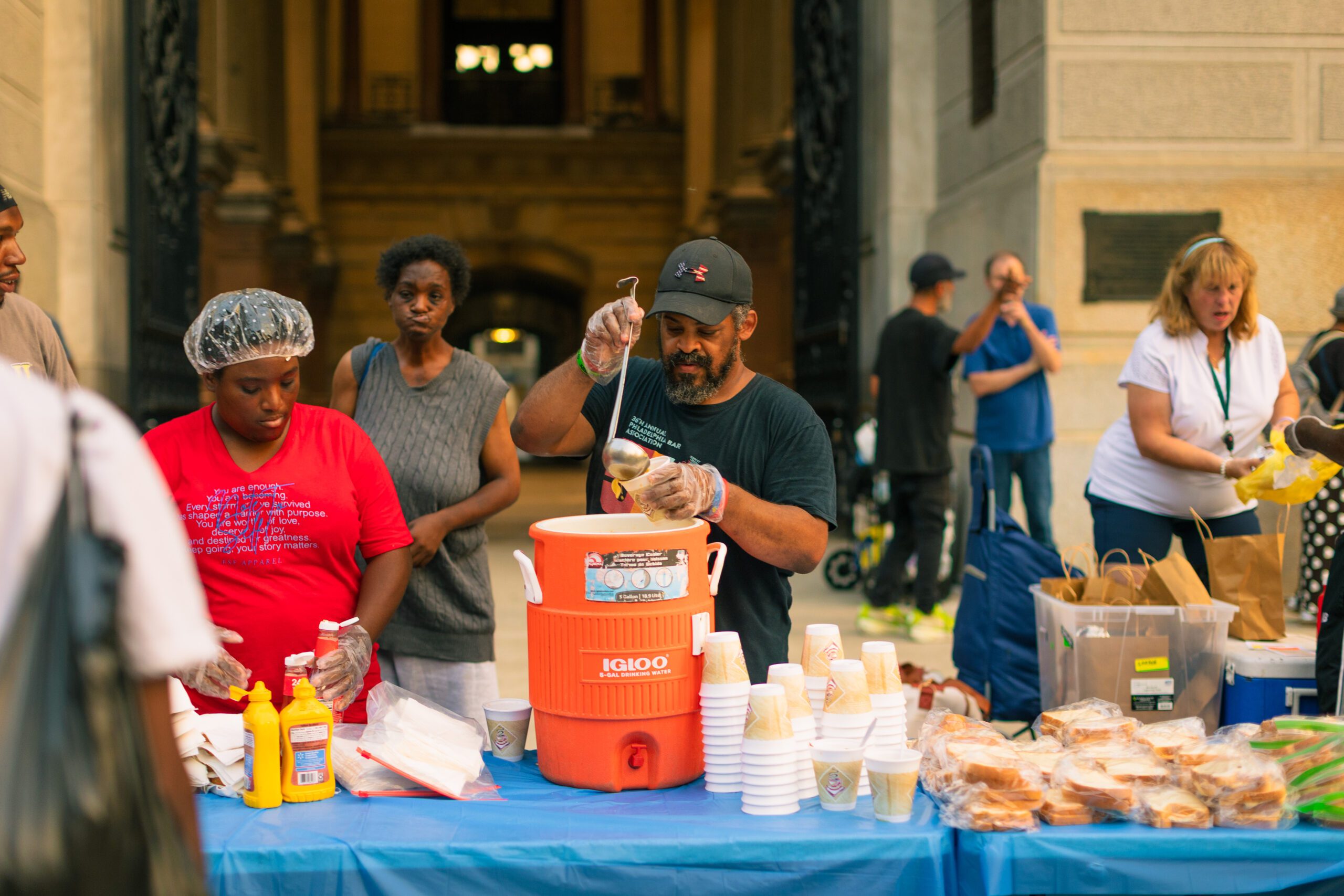 Community volunteers serve food and drinks at a street outreach event, featuring a table with sandwiches, condiments, and cups, while individuals engage in conversation.