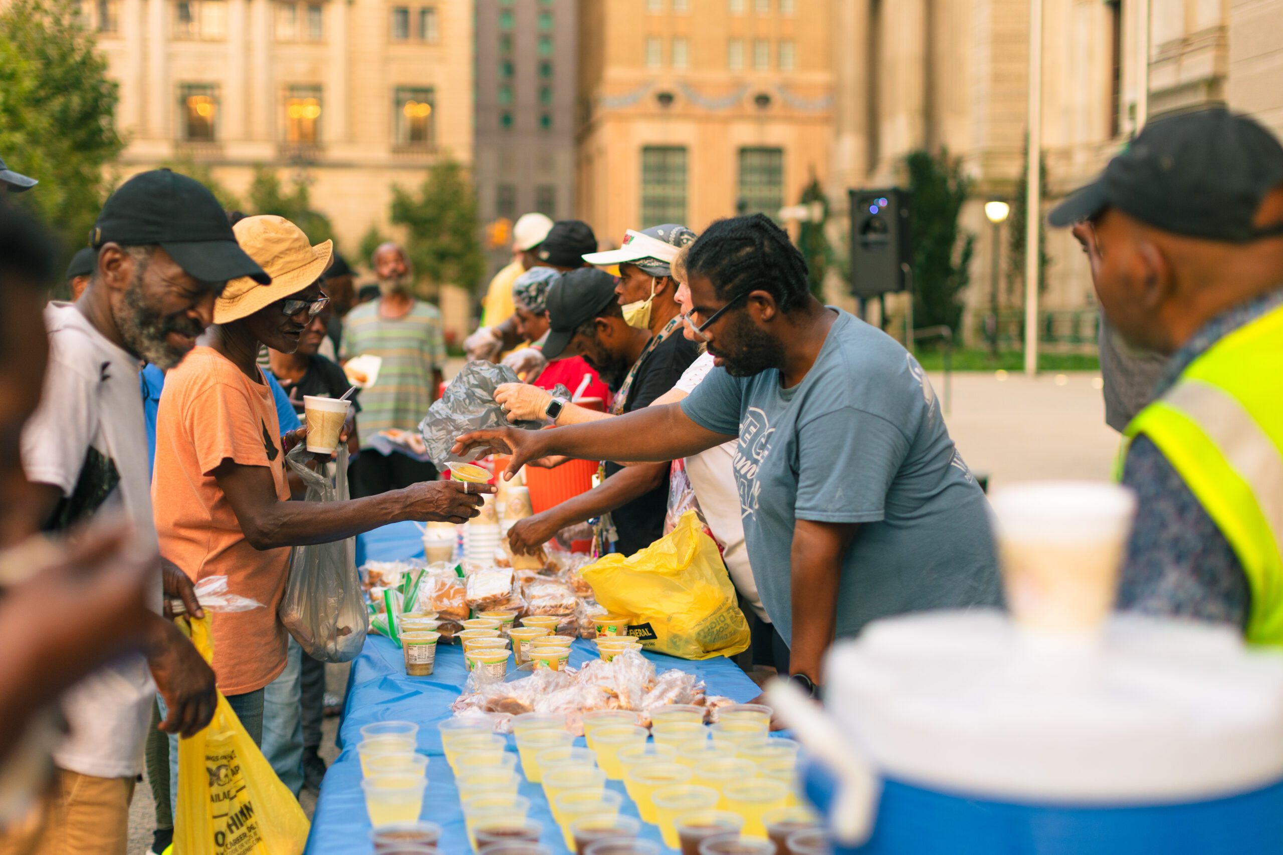 Community members gather at a food distribution event, receiving meals and drinks from volunteers at a table filled with various food items in a public space.