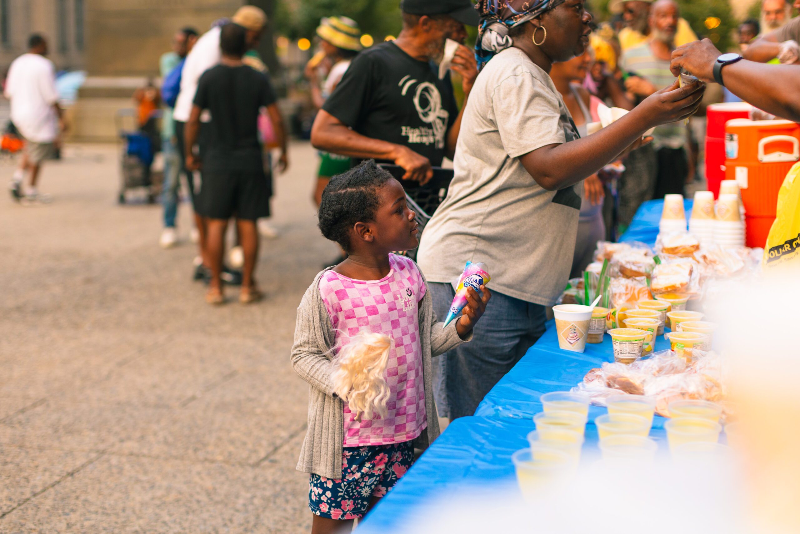 A young girl holding an ice cream cone stands in front of a food table at a community event, surrounded by people enjoying various snacks and beverages in a lively outdoor setting.