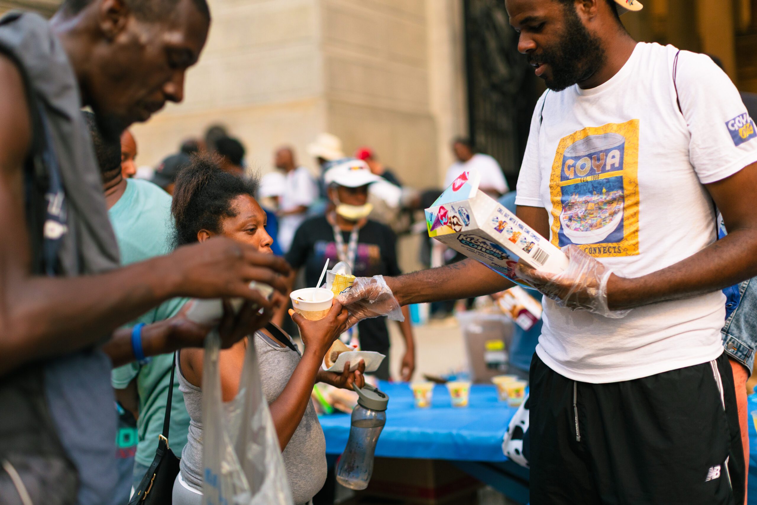 Community members receiving food and drinks at a local outreach event, with a focus on a woman receiving a cup of food from a volunteer wearing gloves. The scene captures the spirit of generosity and support within the community.