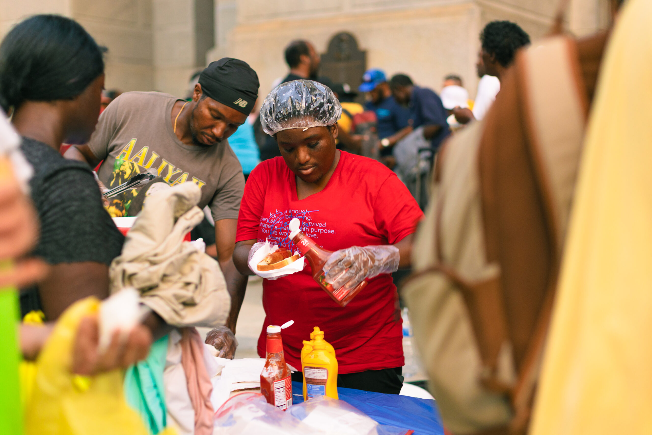 Community volunteers serve food at a local outreach event, with a woman in a red shirt preparing hot dogs and condiments, while attendees gather around in a bustling environment.