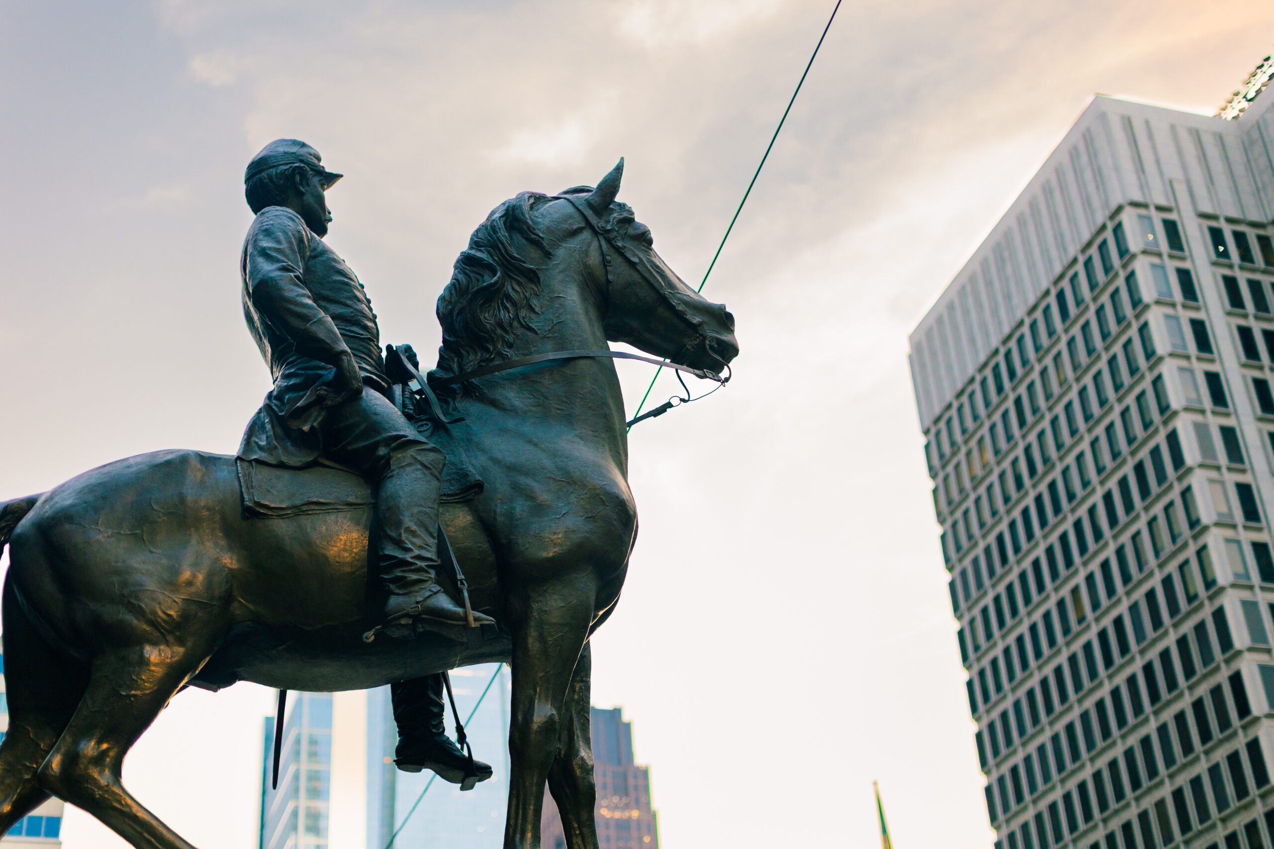 Bronze statue of a mounted soldier, showcasing intricate details of the rider and horse against a backdrop of modern skyscrapers and a cloudy sky.