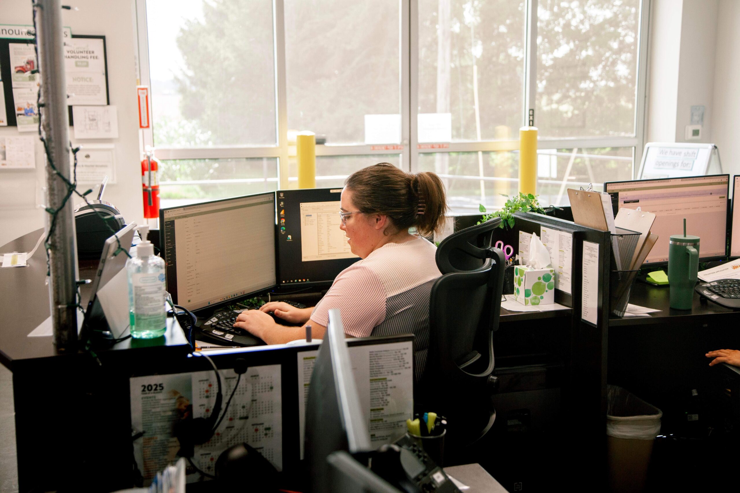 Woman working at a desk with dual monitors in a modern office setting, surrounded by office supplies and plants, focused on her computer tasks.