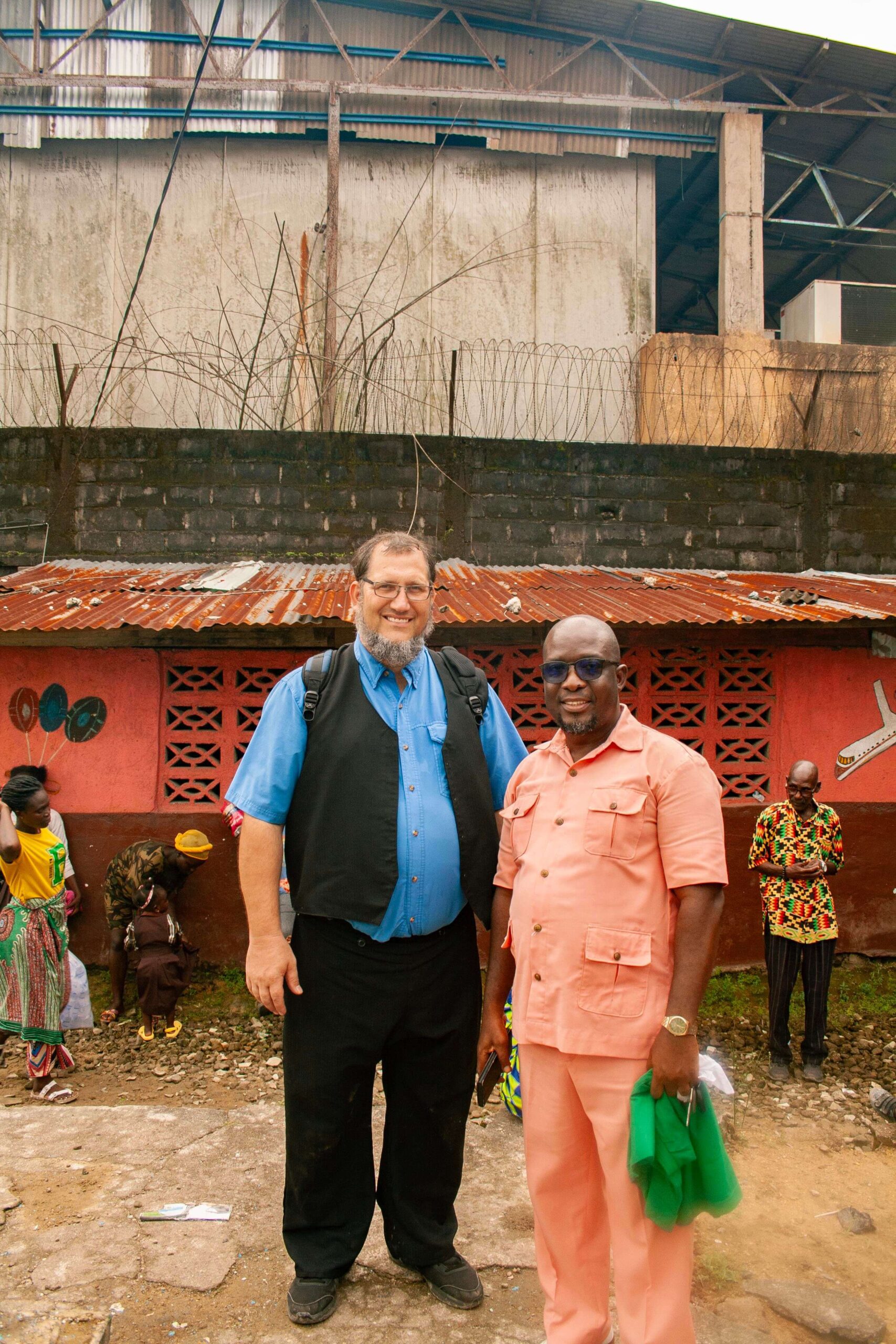 Two men pose for a photo in front of a colorful building in a bustling outdoor market. The man on the left wears a blue shirt and black vest, while the man on the right is dressed in a light pink outfit. In the background, people in traditional clothing are seen engaged in various activities, highlighting the vibrant culture of the area.