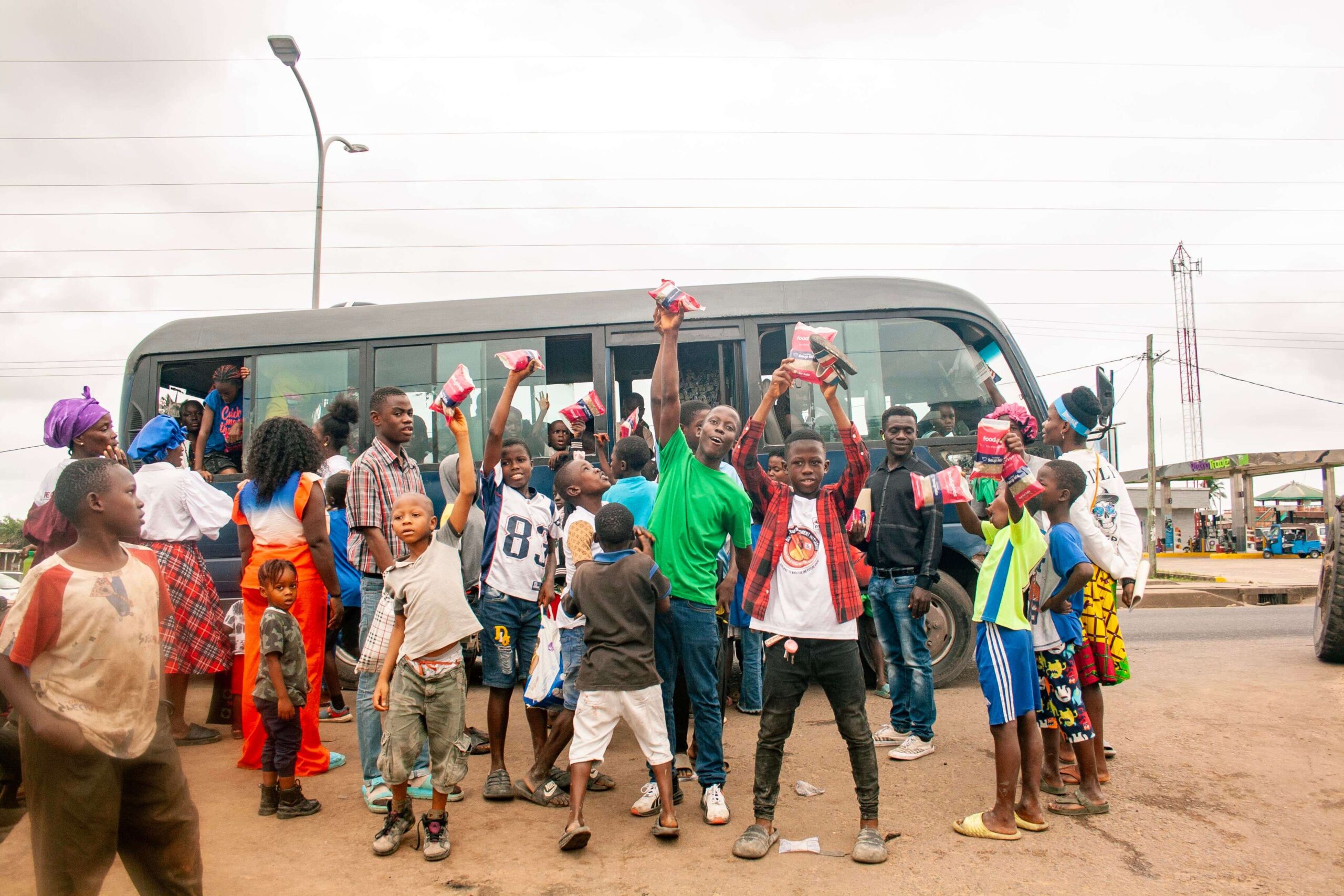 A lively scene of children and adults gathered around a bus, with many children joyfully holding up packages. The image captures a sense of community and excitement, showcasing the interaction between people and the bus, set against a cloudy sky and urban backdrop.