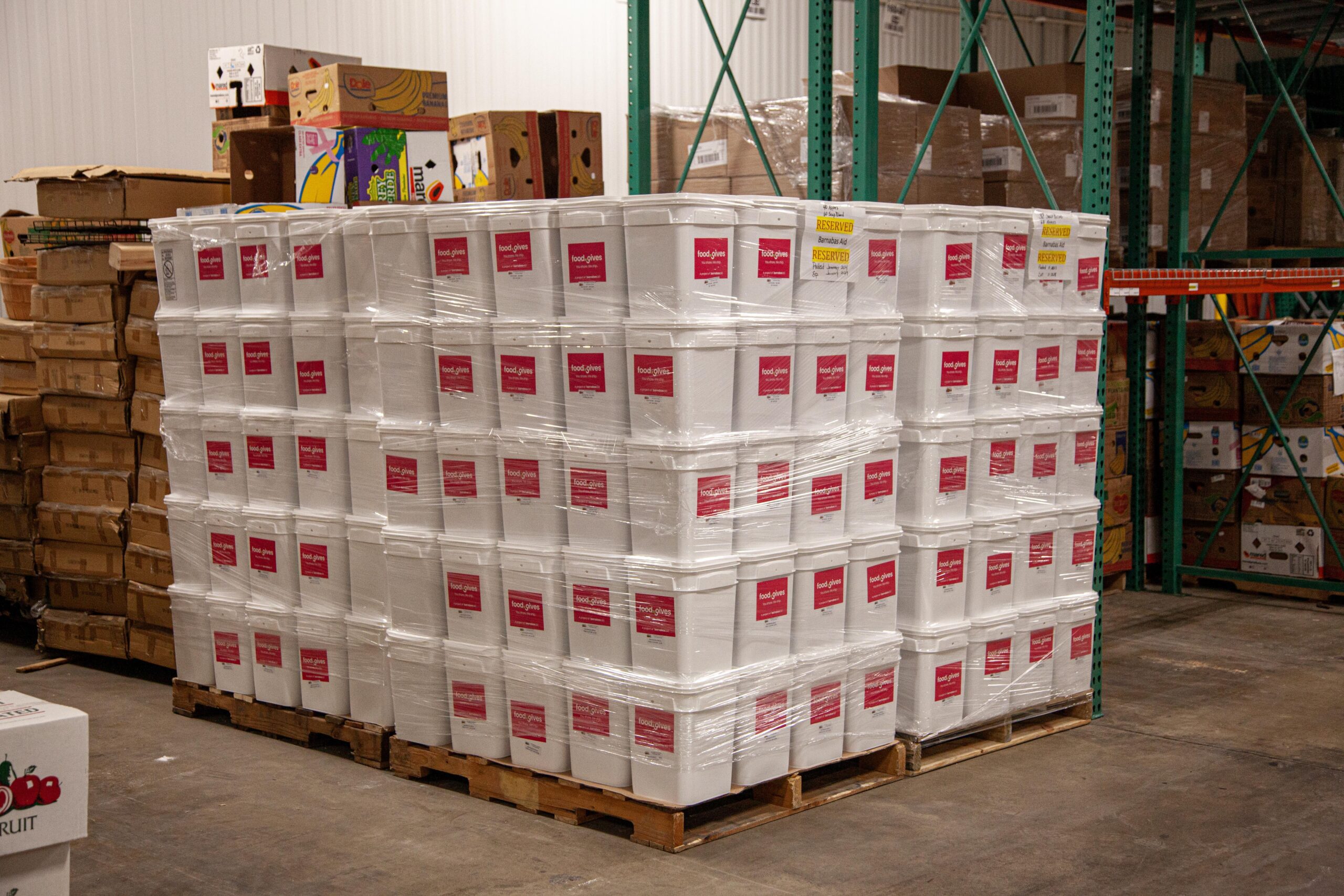 Pallets stacked with white storage bins labeled "food gives" in a warehouse, surrounded by cardboard boxes, showcasing food distribution efforts.