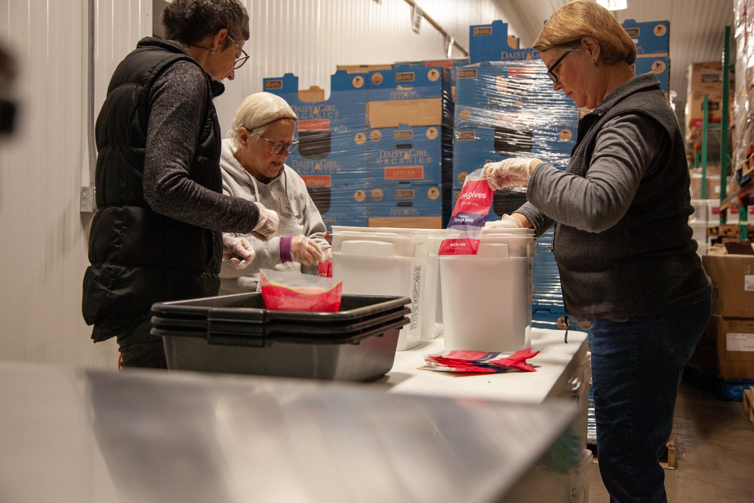 Volunteers preparing food packages in a warehouse, organizing soup mix into containers while wearing gloves for safety and hygiene. Stacks of food boxes are visible in the background, highlighting community support efforts.