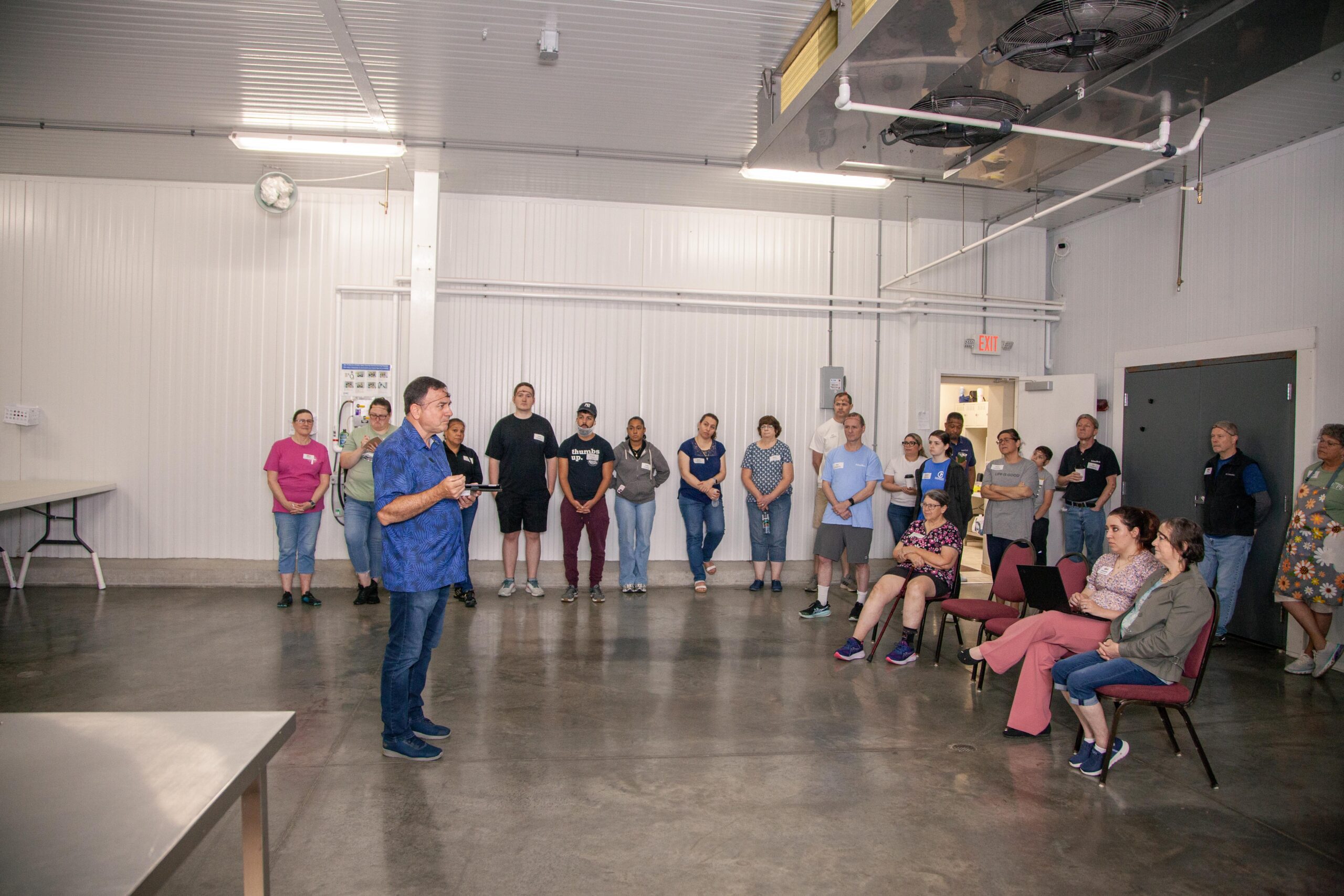 A group of diverse individuals attentively listening to a speaker during a workshop in a well-lit indoor space. The speaker, dressed in a blue shirt, stands in front of the audience, which includes men and women of various ages. Tables and chairs are arranged in the background, emphasizing a collaborative learning environment.