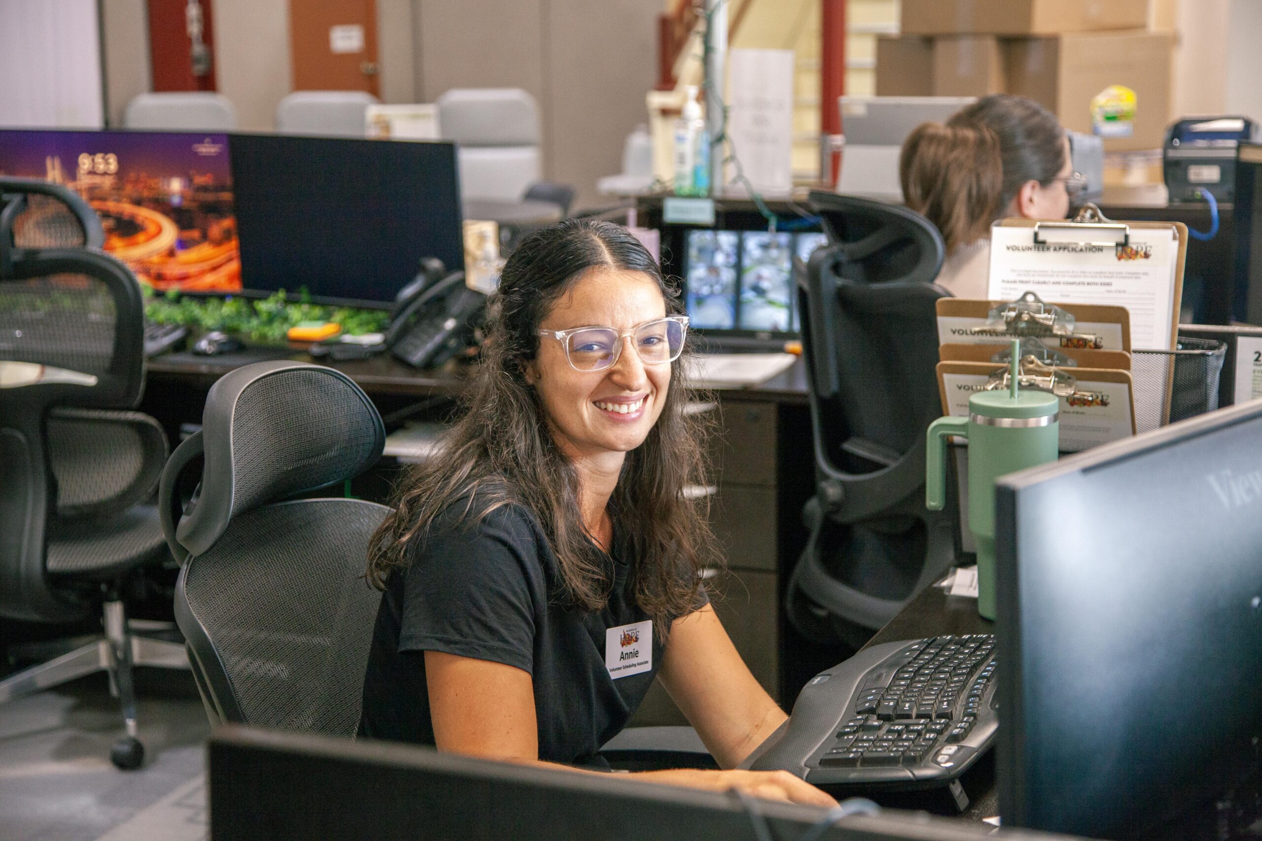 Smiling woman sitting at a desk in an office environment, working on a computer with multiple screens, surrounded by office supplies and paperwork, showcasing a friendly and productive workspace.