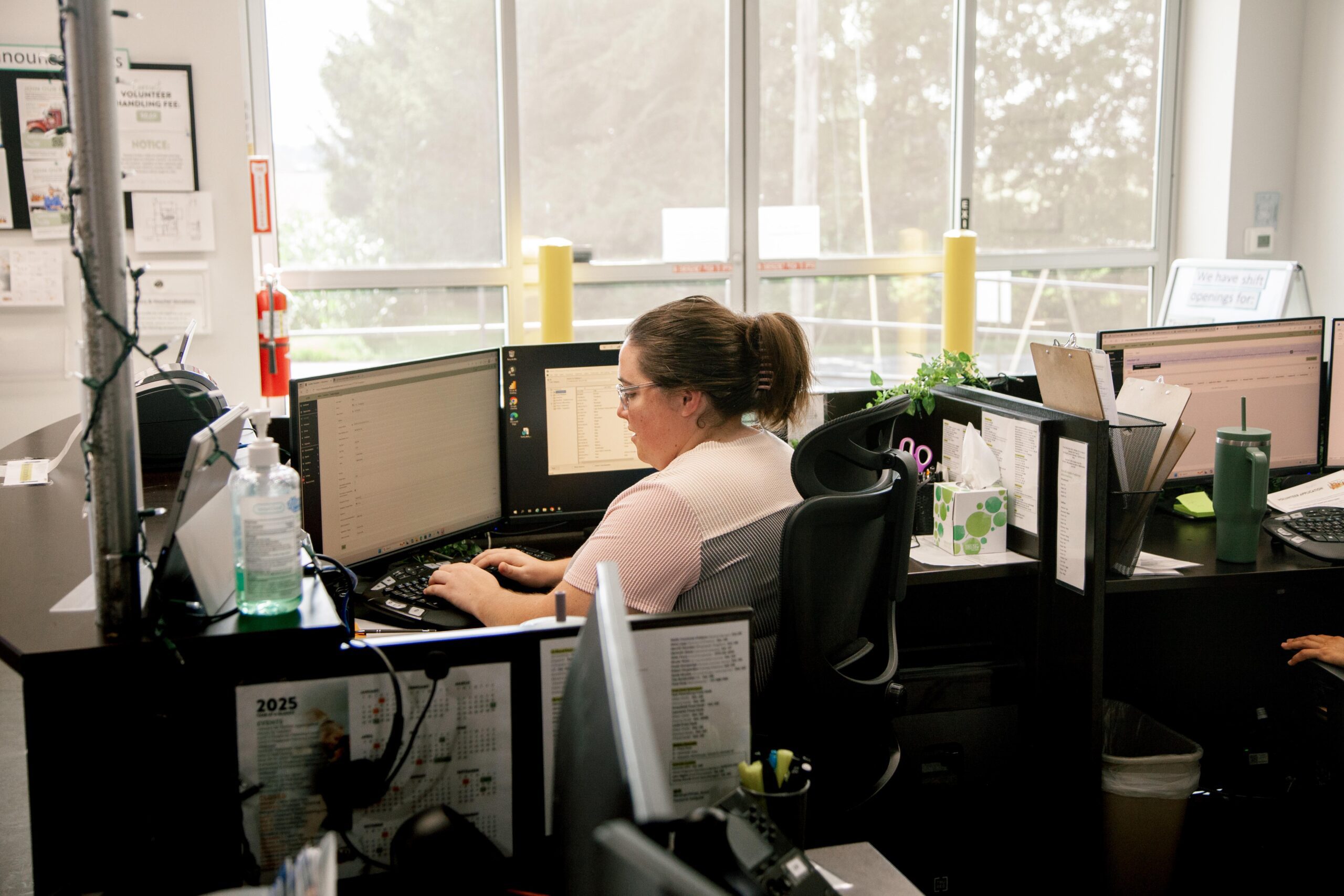 Woman working at a desk with dual monitors in a modern office environment, surrounded by plants and office supplies, focusing on her computer tasks.