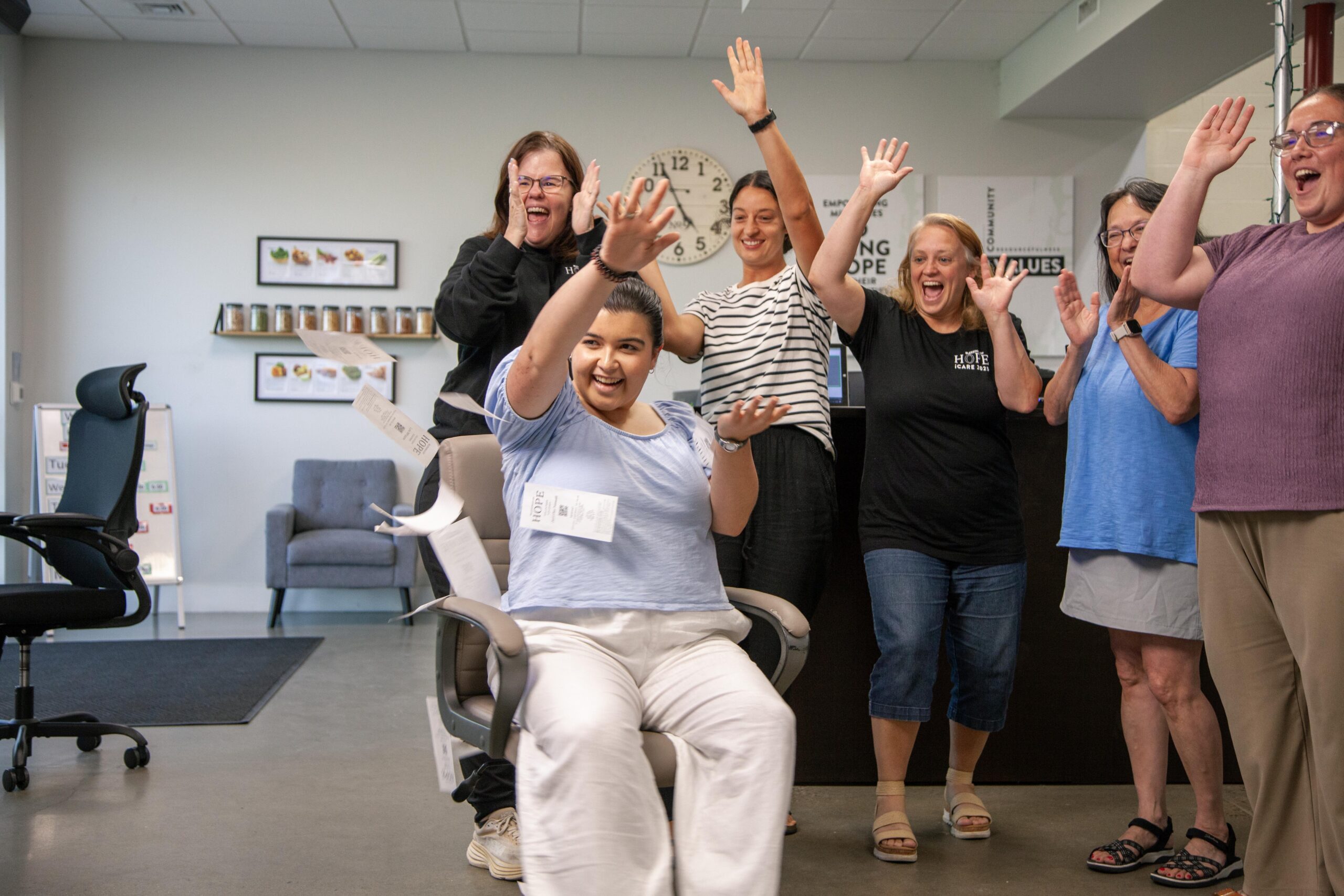 Group of cheerful individuals celebrating in an office setting, with a young woman in a chair surrounded by colleagues raising their hands joyfully. Papers are playfully scattered in the air, showcasing a lively atmosphere of teamwork and positivity.