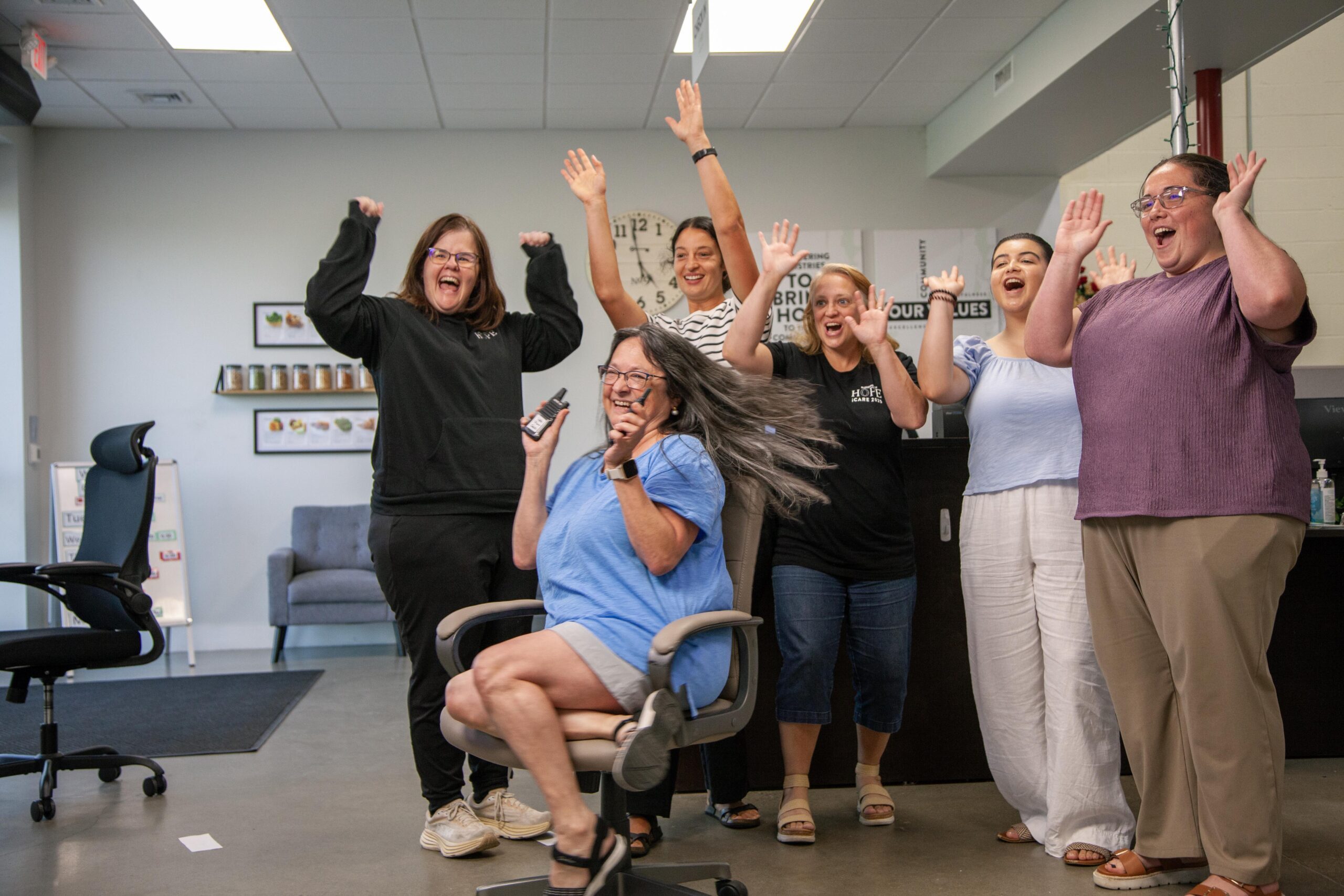 Group of six women celebrating in an office environment, with one woman spinning in a chair and others raising their hands joyfully. The backdrop features a wall with motivational quotes and a shelf displaying jars. The atmosphere is lively and energetic, showcasing teamwork and camaraderie.