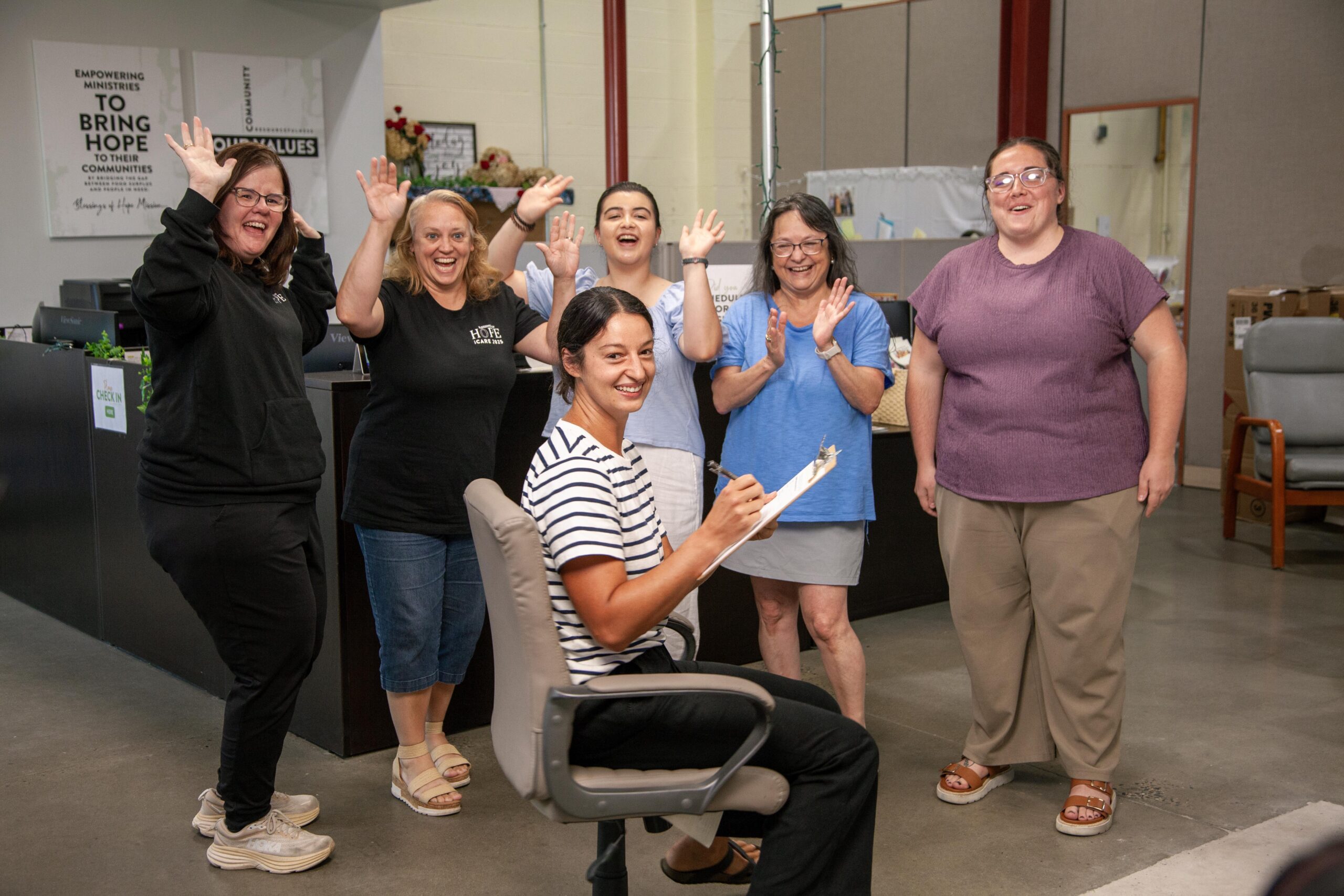 Group of six women smiling and celebrating in an office setting, with one woman seated at a desk writing on a clipboard. The background features signs promoting community empowerment and hope. The atmosphere is joyful and supportive, highlighting teamwork and collaboration.