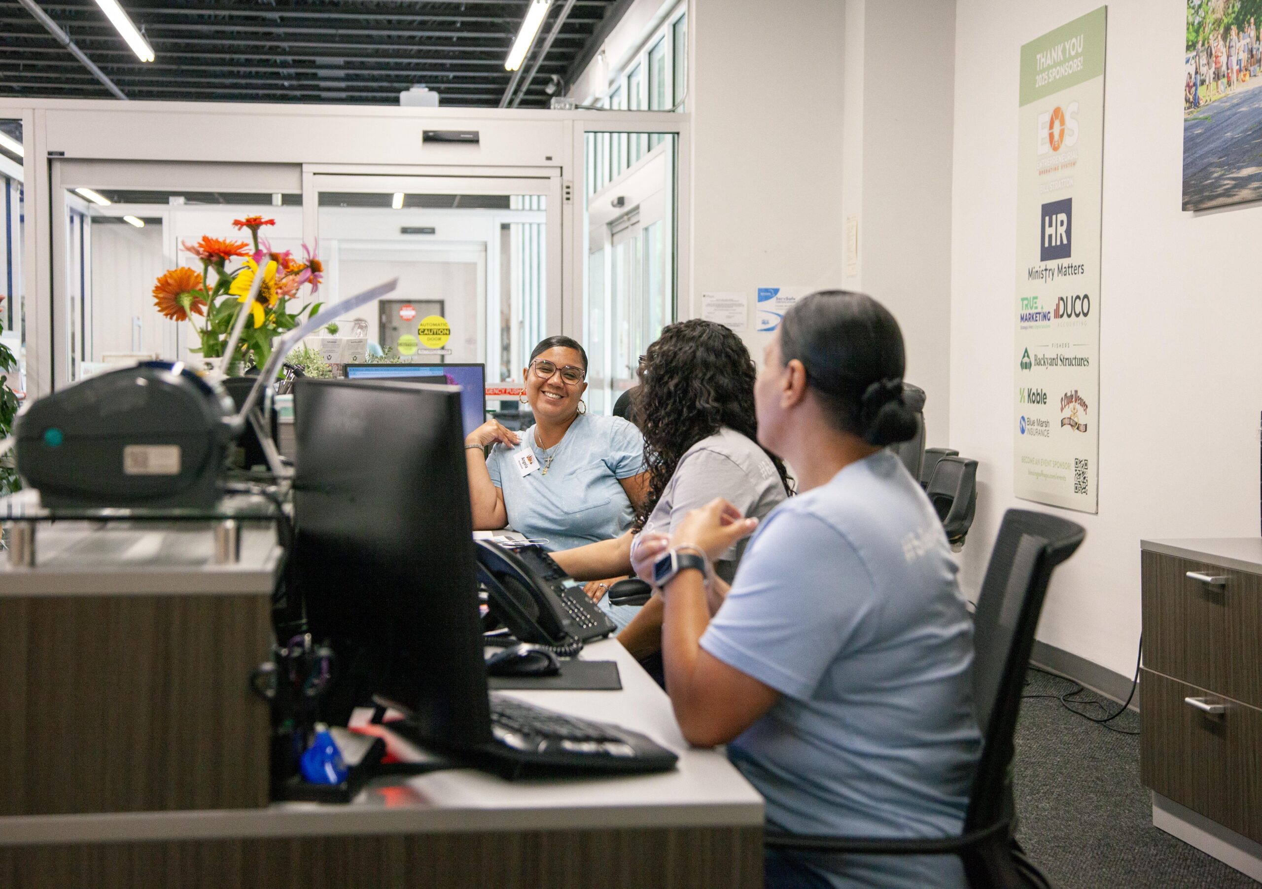 Friendly reception area with three women in light blue shirts engaged in conversation, surrounded by flowers and office equipment. The background features a welcoming entrance and a wall displaying sponsor logos, highlighting a vibrant and community-oriented atmosphere.