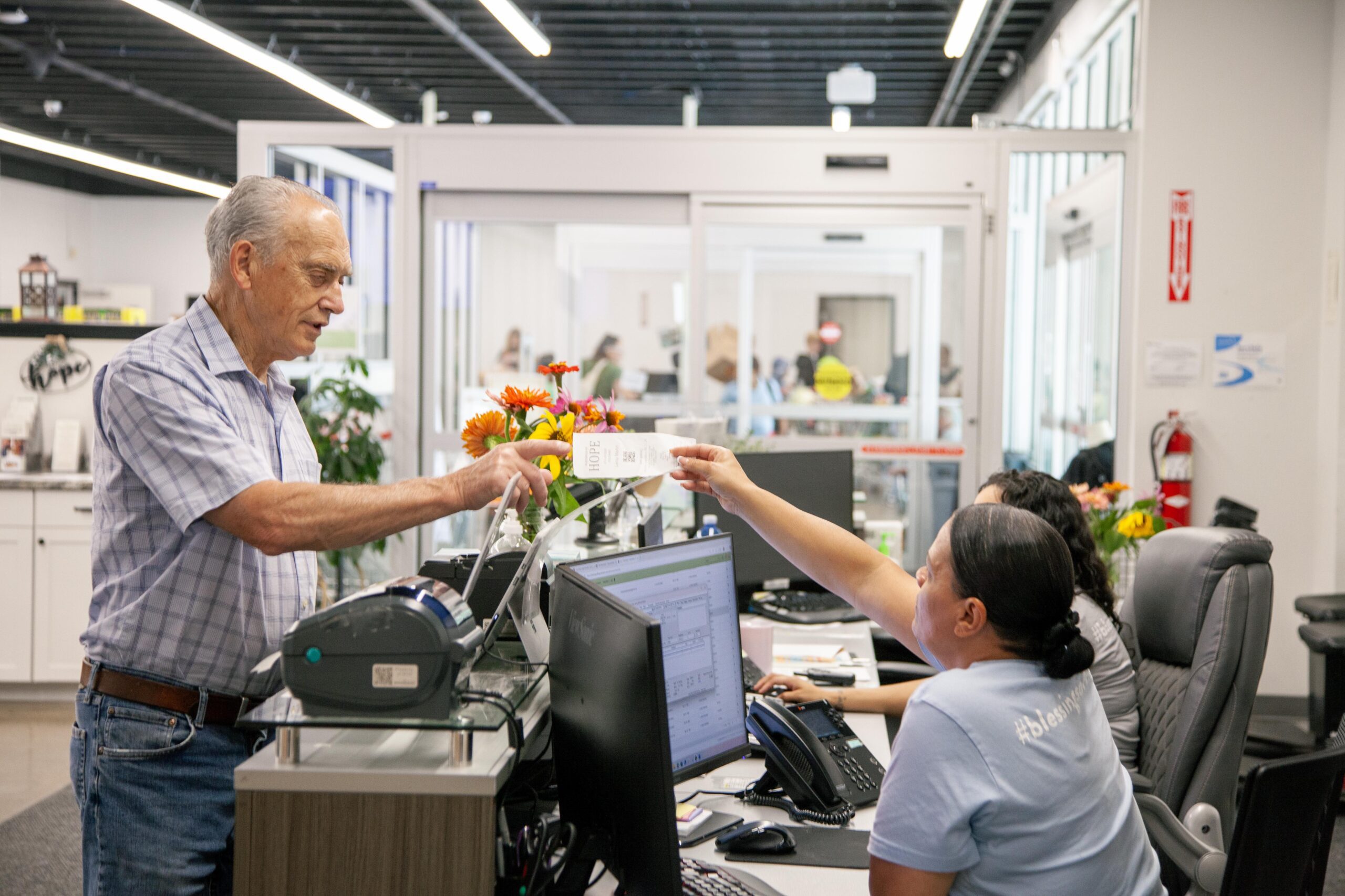 An elderly man handing a receipt to a receptionist at a busy service desk, with flowers and computers visible in the background, highlighting a friendly customer service interaction.