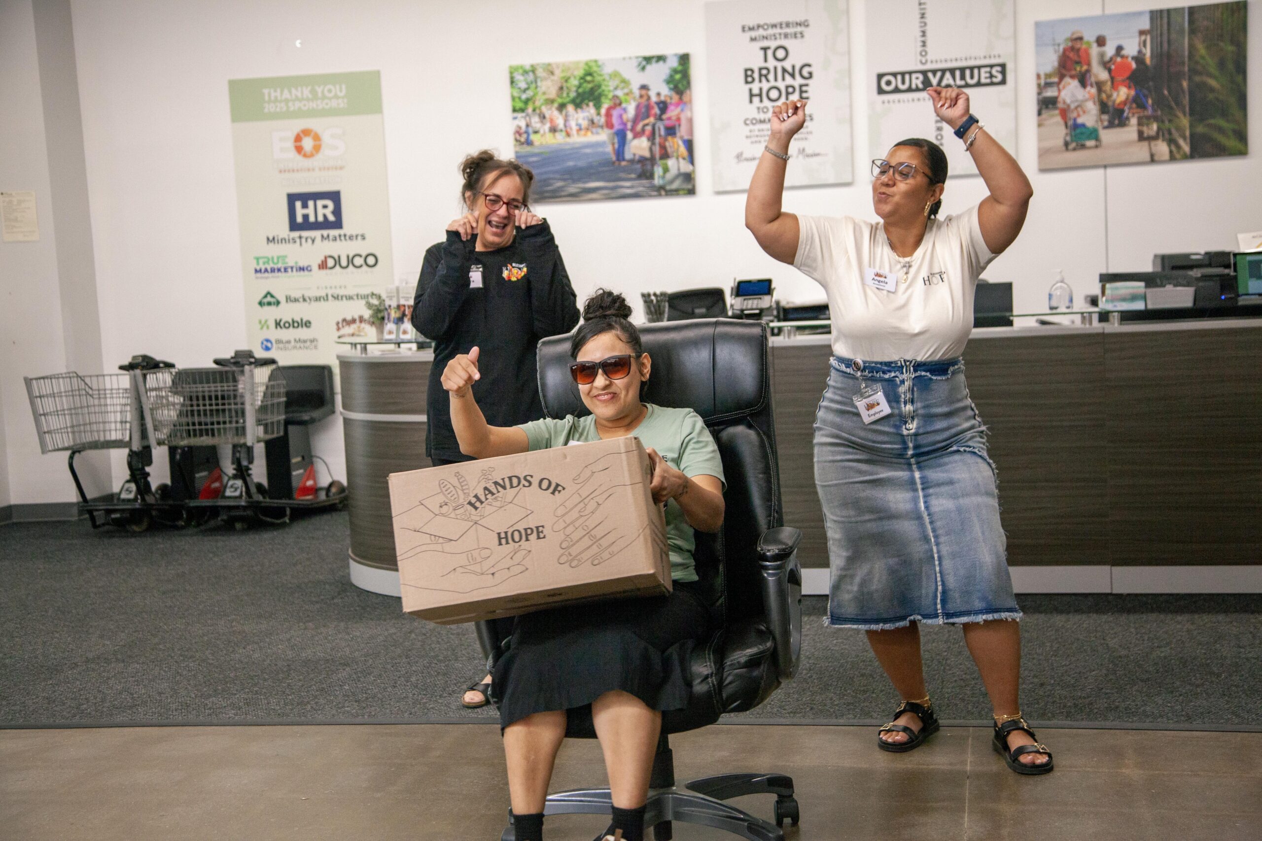 Three women celebrate in an office environment, with one woman sitting in a chair holding a box labeled "Hands of Hope." The other two women are cheering enthusiastically, showcasing a joyful atmosphere. The backdrop includes sponsor logos and motivational signage, emphasizing community support and empowerment.