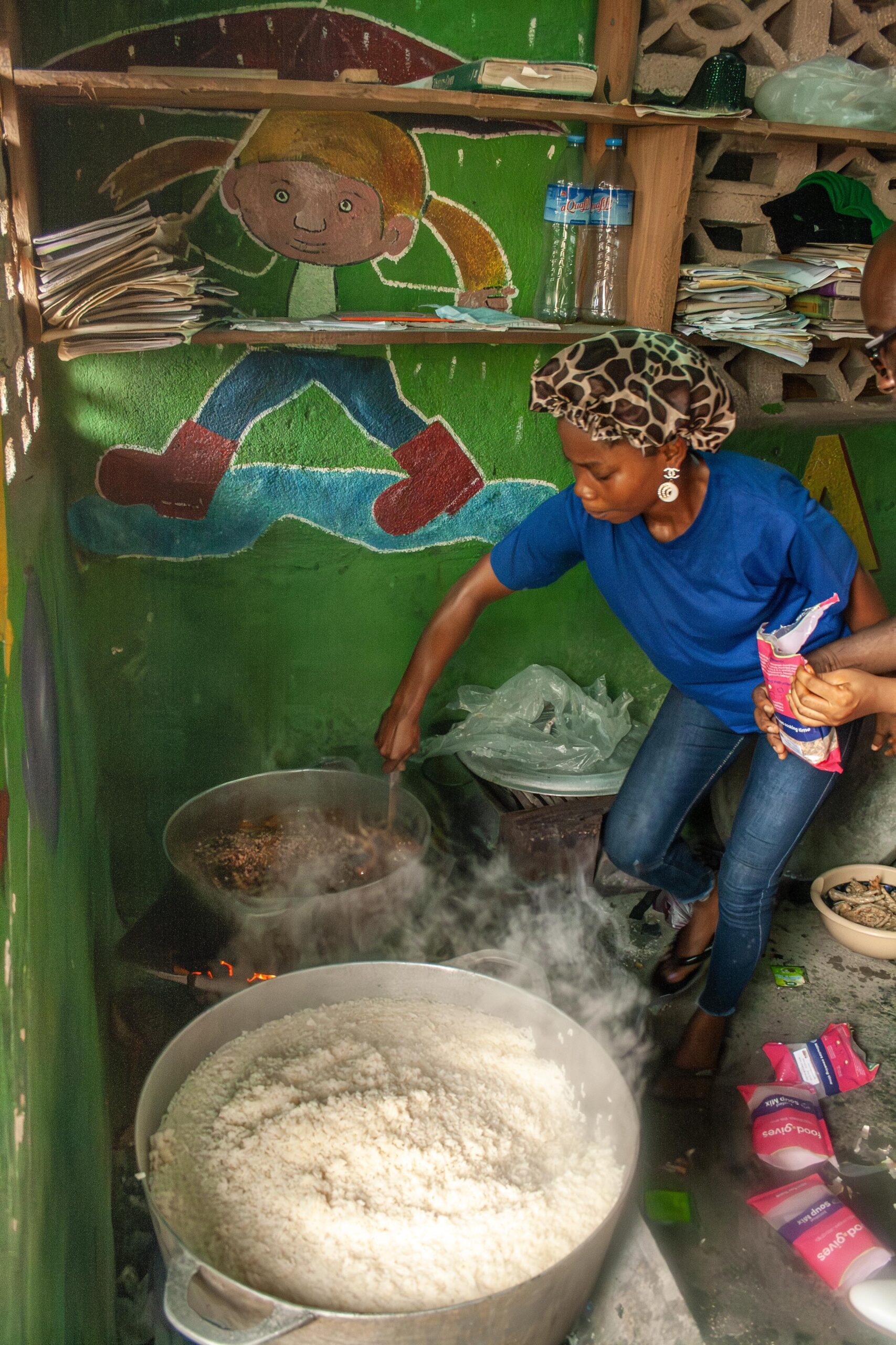 A woman in a blue shirt stirs a large pot of rice while cooking in a colorful kitchen with a mural of a child on the wall. She is surrounded by pots and cooking supplies, with steam rising from the food.